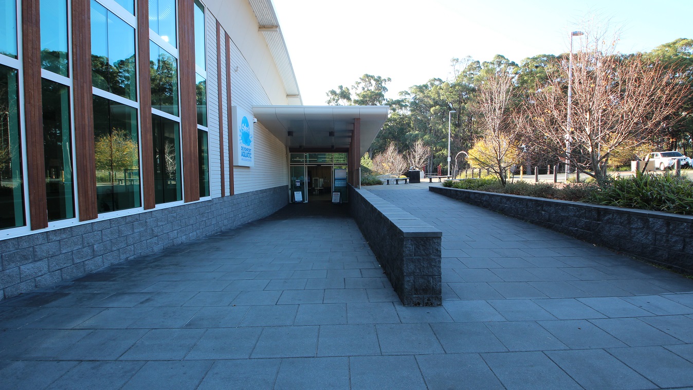 Accessible entrance ramp leading to the main entrance glass doors at Splash Devonport. Rectangular signboards are placed on the glass doors. On the left side wall is the colorful Splash Devonport Aquatic logo. Overlooking this section are tall trees with wide canopies that provide significant shade to the parking area. Accessible entrance ramp leading to the main entrance glass doors at Splash Devonport. Rectangular signboards are placed on the glass doors. On the left side wall is the colorful Splash Devonport Aquatic logo. Overlooking this section are tall trees with wide canopies that provide significant shade to the parking area.