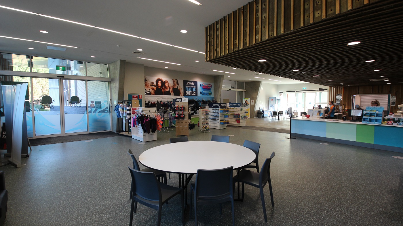 The seating area near the reception is composed of white circular tables with black plastic chairs. Aside from the swimming attire in the retail section are slippers and other swimming equipment. The reception desk is painted in different hues of blue and green color and is run by staff wearing orange and blue polo shirts. The seating area near the reception is composed of white circular tables with black plastic chairs. Aside from the swimming attire in the retail section are slippers and other swimming equipment. The reception desk is painted in different hues of blue and green color and is run by staff wearing orange and blue polo shirts.