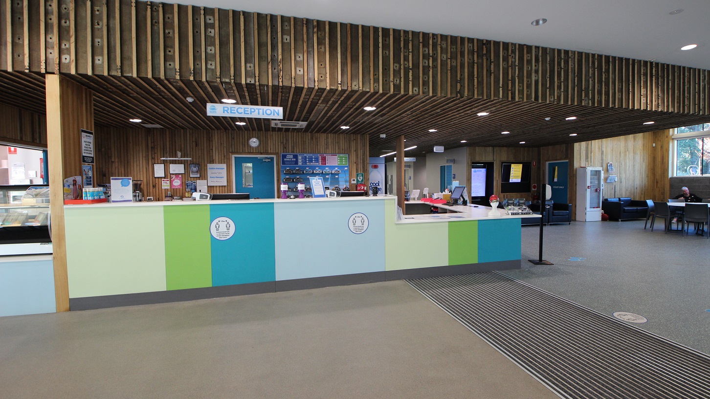 The reception area can be distinguished by the word reception hung on the wooden ceiling and a pattern of parallel lines in front of the reception area. The reception countertop is painted in different hues of green and blue. On the left side is the café, while on the right side is a liquid soap and alcohol dispenser in the reception area. The reception area can be distinguished by the word reception hung on the wooden ceiling and a pattern of parallel lines in front of the reception area. The reception countertop is painted in different hues of green and blue. On the left side is the café, while on the right side is a liquid soap and alcohol dispenser in the reception area.