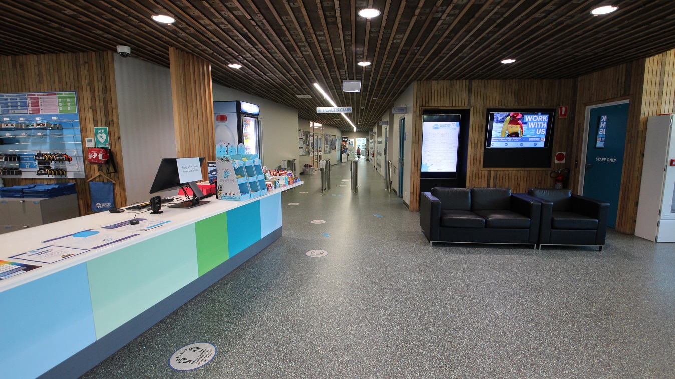 Heading straight from the main entrance doors and past the reception area is the corridor leading to the Health Club. The entry point to the Health Club is by the grey metal turnstiles. Opposite the reception desk are a staff-only blue-green door, black sofa chairs, and a television mounted on the wooden walls. Heading straight from the main entrance doors and past the reception area is the corridor leading to the Health Club. The entry point to the Health Club is by the grey metal turnstiles. Opposite the reception desk are a staff-only blue-green door, black sofa chairs, and a television mounted on the wooden walls.
