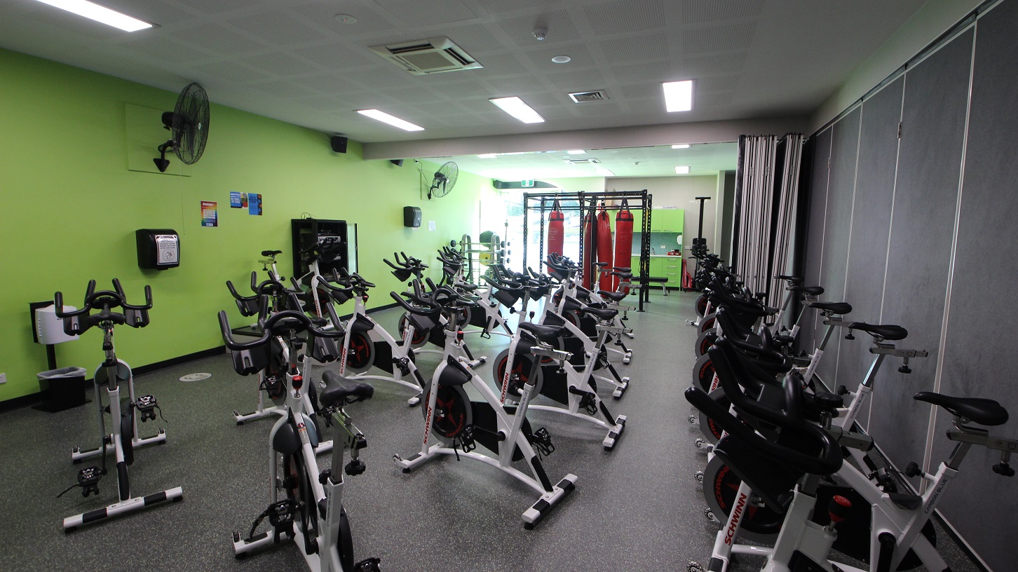 Inside the cycle room at Splash Devonport. Several stationary bikes of grey, black, and white are arranged in three rows and are available for use. Movable walls painted in grey at the back separate the cycle room from the other. The other walls are painted neon green and a large, black, wall fans are mounted on the wall for ventilation. Four red punching bags are also hung on a black metal frame. Inside the cycle room at Splash Devonport. Several stationary bikes of grey, black, and white are arranged in three rows and are available for use. Movable walls painted in grey at the back separate the cycle room from the other. The other walls are painted neon green and a large, black, wall fans are mounted on the wall for ventilation. Four red punching bags are also hung on a black metal frame.