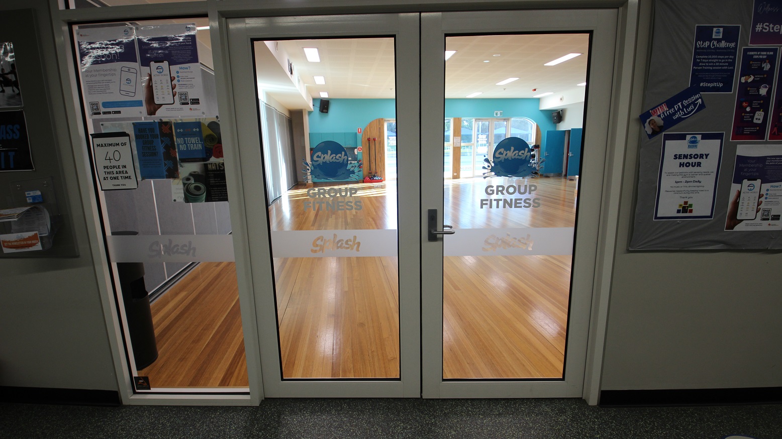 Manually operated glass double door of Group Fitness room at Splash Devonport. At the entrance door is a paper telling guests that only 40 people can utilize the facility at one time. Seen through the glass doors are the brown walnut wood flooring and the sky-blue wall and another glass door overlooking the outside pool. At the back of the room is the movable grey wall. Manually operated glass double door of Group Fitness room at Splash Devonport. At the entrance door is a paper telling guests that only 40 people can utilize the facility at one time. Seen through the glass doors are the brown walnut wood flooring and the sky-blue wall and another glass door overlooking the outside pool. At the back of the room is the movable grey wall.