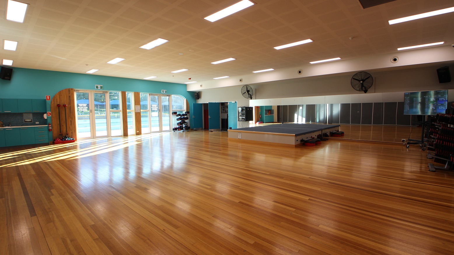 Large open space inside the Group Fitness gymnasium. The brown walnut wooden flooring shines beautifully as light strikes through it. Large mirrors are placed on the wall in front. A stage with a grey top that leaders could step into during fitness dance. Wall fans are mounted on the front wall of the room for proper ventilation and two blue doors on the left side are open. Large open space inside the Group Fitness gymnasium. The brown walnut wooden flooring shines beautifully as light strikes through it. Large mirrors are placed on the wall in front. A stage with a grey top that leaders could step into during fitness dance. Wall fans are mounted on the front wall of the room for proper ventilation and two blue doors on the left side are open.