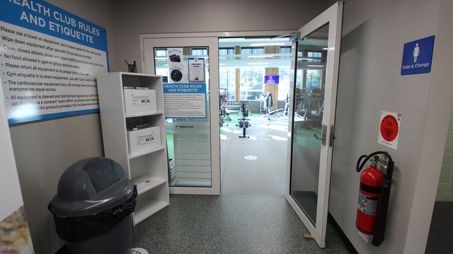 The other half of the manually operated double doors of Health Club at Splash Devonport is open while the other is not. Health Club rules and etiquette can be found on the doors and walls in small and large fonts for everyone to read. There is a large, grey, and black trash bin, a white cabinet, and a fire distinguisher at the entrance doors. The other half of the manually operated double doors of Health Club at Splash Devonport is open while the other is not. Health Club rules and etiquette can be found on the doors and walls in small and large fonts for everyone to read. There is a large, grey, and black trash bin, a white cabinet, and a fire distinguisher at the entrance doors.