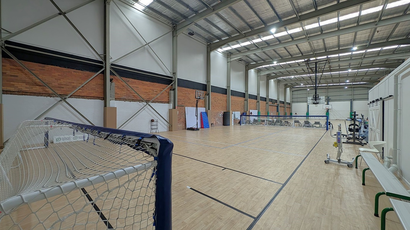 A spacious indoor sports hall with a wooden floor and high metal ceiling. On the left, a goal net is visible. The walls are a mix of brick and white panels with metal supports. In the centre, a basketball hoop and a badminton net are set up. On the right, there are gym equipment, benches, and chairs lined up along the wall. The ceiling has large fluorescent lights.