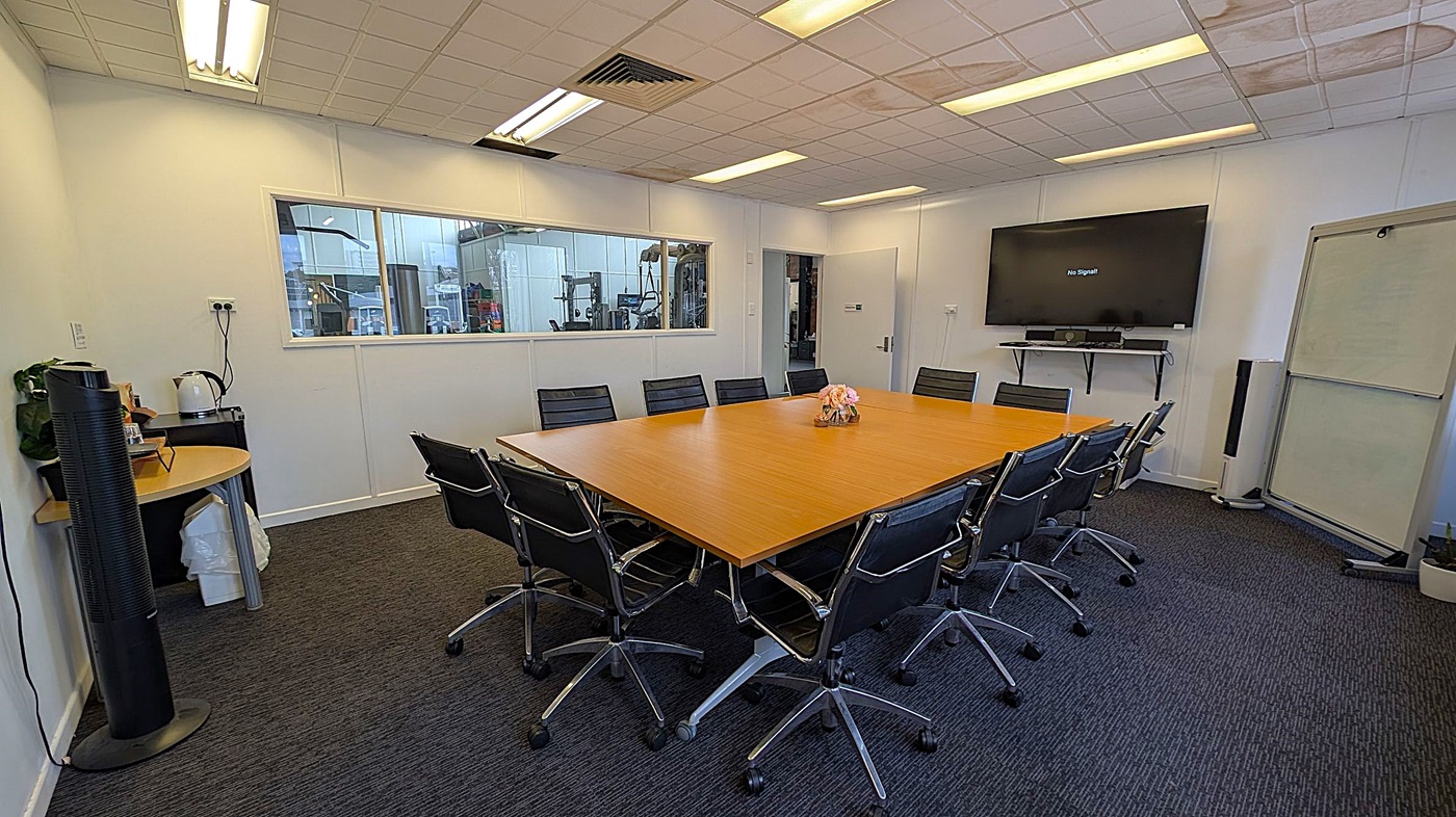A conference room with a large rectangular wooden table surrounded by black office chairs on wheels. On the right wall, a large flat-screen TV displays "No Signal" above a shelf with a soundbar. A whiteboard stands nearby. On the left, a small round table holds a kettle and other items. A window in the back wall reveals an industrial area. The ceiling has fluorescent lights, and the floor is carpeted in dark grey.
