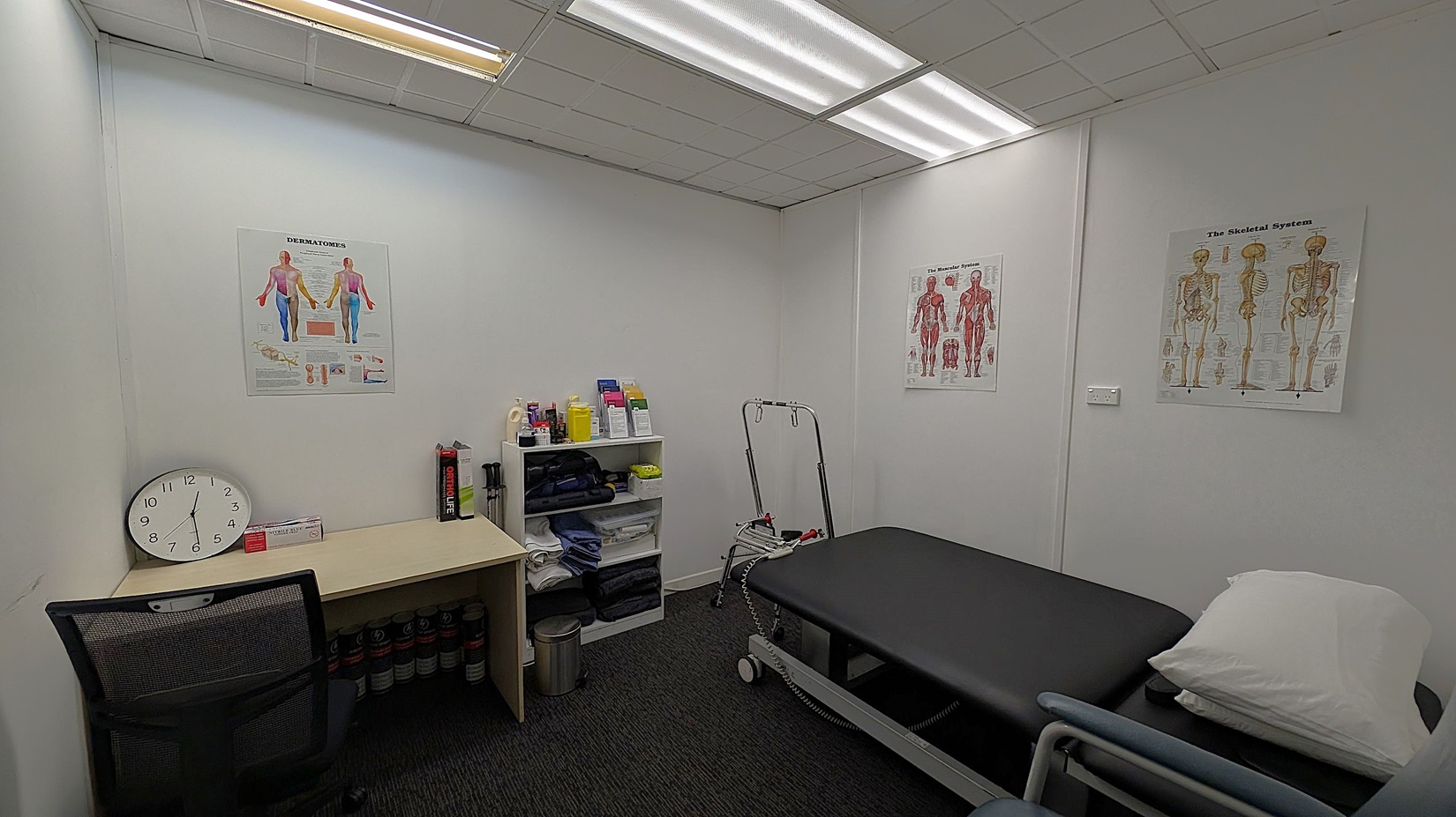 A small medical examination room with white walls and a dark carpeted floor. On the left, there's a desk with a large clock, a chair, and some medical supplies. In the centre, a shelving unit holds various bottles and pamphlets. On the right, a black examination table with a white pillow is positioned. The walls display anatomical posters. The ceiling has fluorescent lighting.