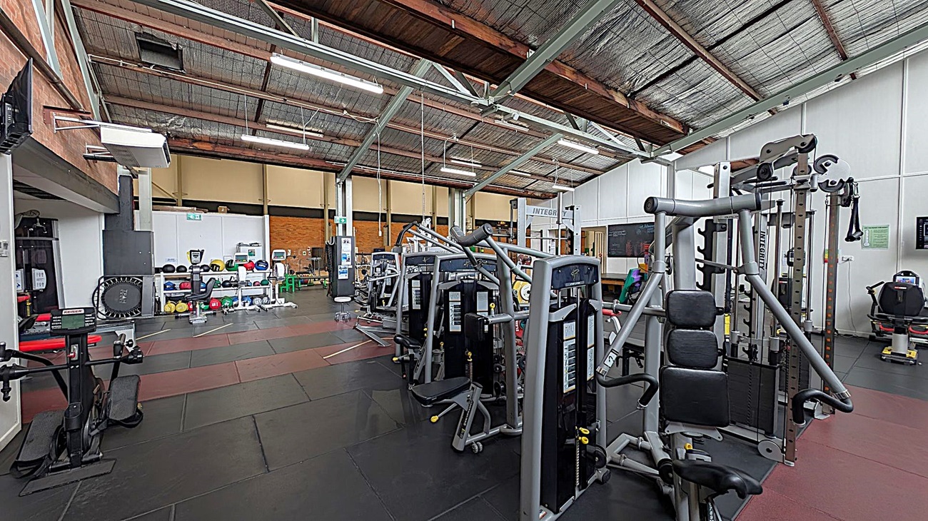 A gym interior with various exercise machines. On the right, there are weight machines and a leg press. In the middle, more weight machines are aligned. On the left, a trampoline and shelves with medicine balls and kettlebells are visible. The floor is covered with black and red mats, and the ceiling has exposed beams and lighting fixtures. The walls are a mix of brick and white panels.