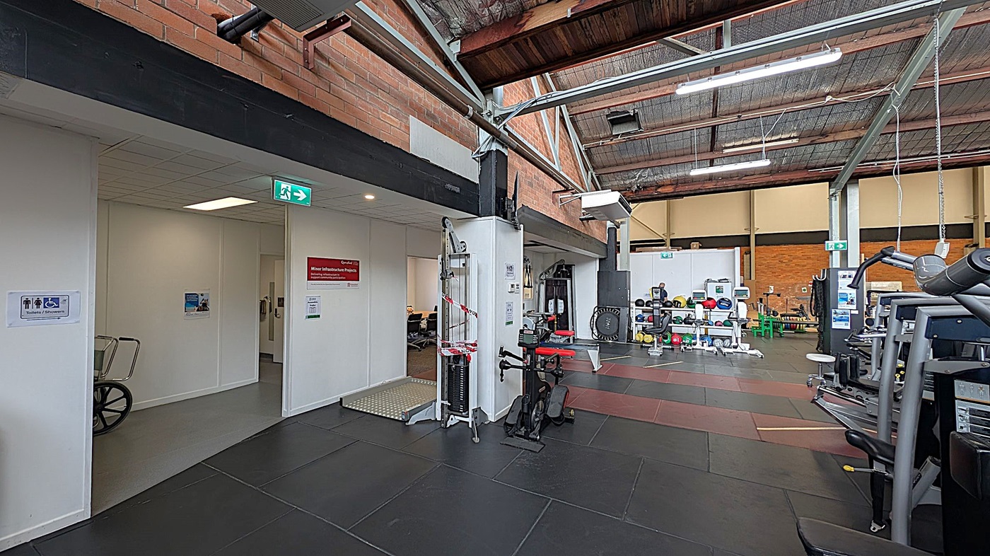 A gym interior with a high, industrial-style ceiling. On the left, there's a doorway leading to a hallway with a wheelchair sign and an exit sign above. A piece of gym equipment is partially visible near the entrance. In the centre, various gym machines and weights are arranged, including a cable machine with red and white tape. On the right, more gym equipment is visible, including a treadmill. The floor is covered with black and brown rubber mats, and the walls are brick.