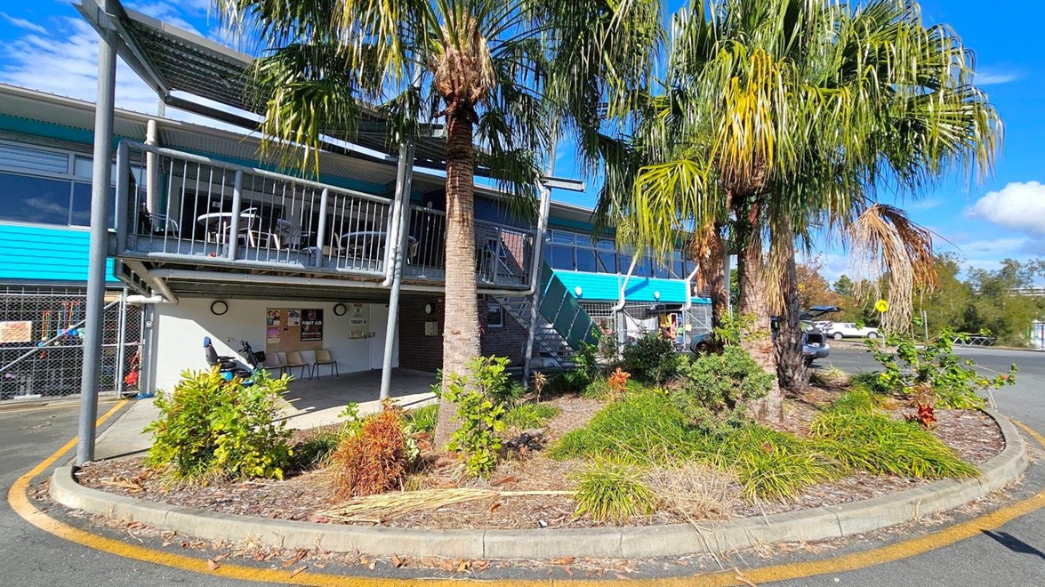 A building with a blue and grey exterior, featuring a balcony with metal railings and outdoor seating on the left. Below the balcony, there are posters and a first aid sign on the wall. In the foreground, a landscaped area with palm trees and various shrubs is surrounded by a kerb. To the right, a car park with a few vehicles is visible, along with a road and more greenery in the background. A building with a blue and grey exterior, featuring a balcony with metal railings and outdoor seating on the left. Below the balcony, there are posters and a first aid sign on the wall. In the foreground, a landscaped area with palm trees and various shrubs is surrounded by a kerb. To the right, a car park with a few vehicles is visible, along with a road and more greenery in the background.