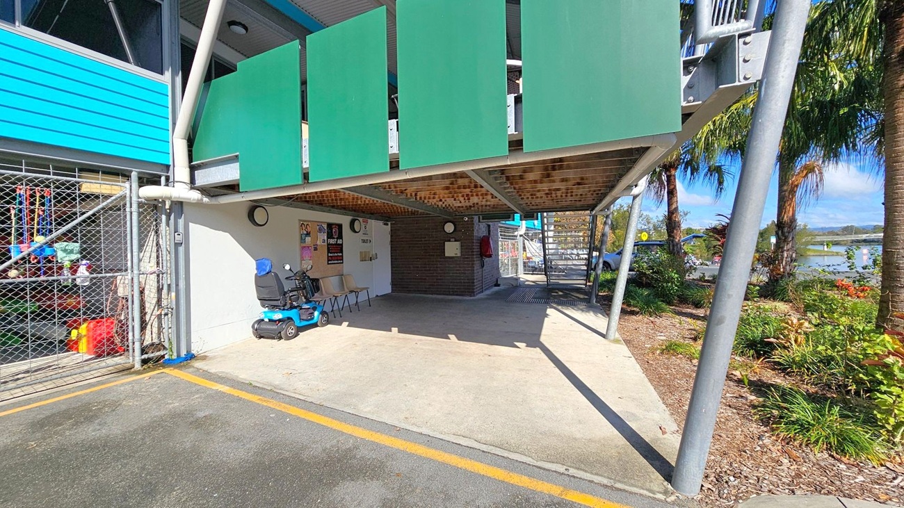 Under a building with green panels, there's a sheltered area with a blue mobility scooter and two chairs. A chain-link fence on the left encloses various items. The ground is a mix of concrete and asphalt with a yellow line. On the right, a garden with palm trees and shrubs is visible. A staircase leads up from the back, and a body of water can be seen in the distance. The ceiling is supported by metal beams. Under a building with green panels, there's a sheltered area with a blue mobility scooter and two chairs. A chain-link fence on the left encloses various items. The ground is a mix of concrete and asphalt with a yellow line. On the right, a garden with palm trees and shrubs is visible. A staircase leads up from the back, and a body of water can be seen in the distance. The ceiling is supported by metal beams.