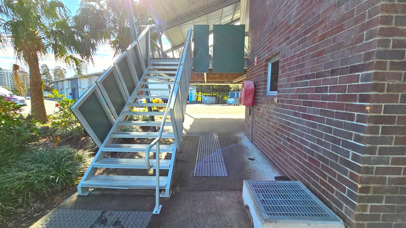 A metal staircase with a handrail on the left leads up to a building. The building has a brick exterior with a small window and a red box mounted on the wall. To the left of the stairs, there are palm trees and a garden area. In the background, there are industrial buildings and a parked car. The ground is concrete with metal grates, and the ceiling of the structure above is metal. A metal staircase with a handrail on the left leads up to a building. The building has a brick exterior with a small window and a red box mounted on the wall. To the left of the stairs, there are palm trees and a garden area. In the background, there are industrial buildings and a parked car. The ground is concrete with metal grates, and the ceiling of the structure above is metal.