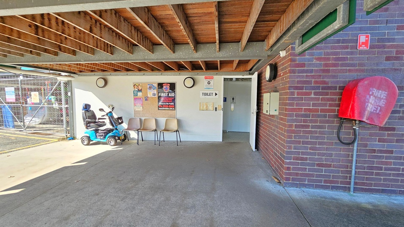 A covered area with a concrete floor and a wooden beam ceiling. On the left, a blue mobility scooter is parked next to three beige chairs. Posters and a clock are mounted on the white wall. A sign points to a toilet through an open door. On the right, a red fire hose reel is attached to a brick wall. In the background, a chain-link fence with various signs is visible. A covered area with a concrete floor and a wooden beam ceiling. On the left, a blue mobility scooter is parked next to three beige chairs. Posters and a clock are mounted on the white wall. A sign points to a toilet through an open door. On the right, a red fire hose reel is attached to a brick wall. In the background, a chain-link fence with various signs is visible.