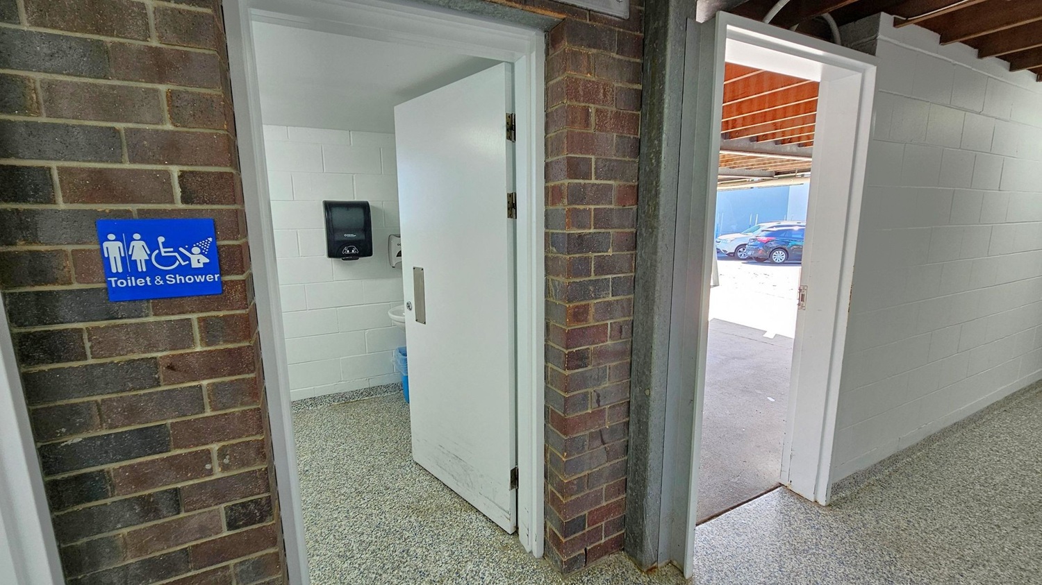 A brick wall on the left features a blue sign indicating a toilet and shower, including accessibility symbols. A white door is partially open, revealing a white-tiled interior with a paper towel dispenser. The floor is speckled grey. On the right, another open door leads outside, where several cars are parked. The ceiling has exposed wooden beams. A brick wall on the left features a blue sign indicating a toilet and shower, including accessibility symbols. A white door is partially open, revealing a white-tiled interior with a paper towel dispenser. The floor is speckled grey. On the right, another open door leads outside, where several cars are parked. The ceiling has exposed wooden beams.
