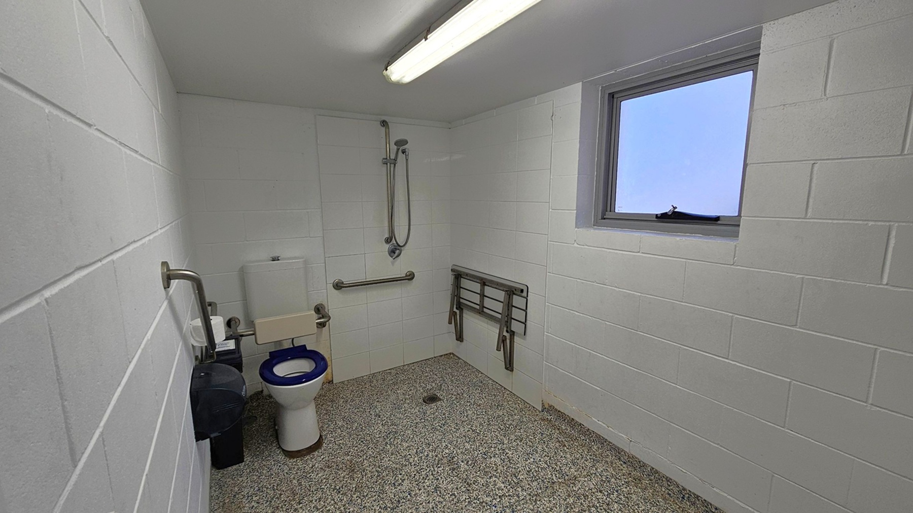 A bathroom with white tiled walls and a speckled grey floor. On the left, there is a toilet with a blue seat, equipped with grab bars and a toilet paper holder. Above the toilet, a cistern is visible. In the centre, a shower area with a handheld showerhead and a horizontal grab bar. On the right, a small window with a handle is set into the wall. A foldable shower seat is mounted on the wall beneath the window. A fluorescent light fixture is on the ceiling. A bathroom with white tiled walls and a speckled grey floor. On the left, there is a toilet with a blue seat, equipped with grab bars and a toilet paper holder. Above the toilet, a cistern is visible. In the centre, a shower area with a handheld showerhead and a horizontal grab bar. On the right, a small window with a handle is set into the wall. A foldable shower seat is mounted on the wall beneath the window. A fluorescent light fixture is on the ceiling.