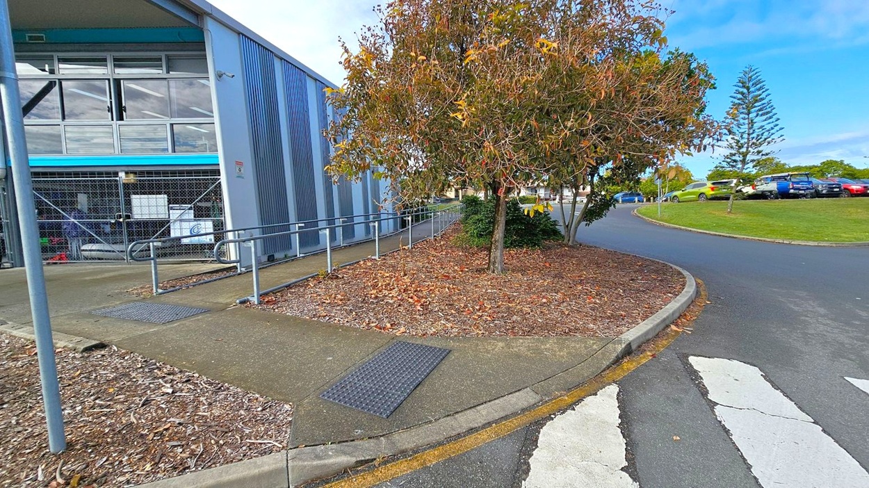 On the left, a building with metal siding and large windows, featuring a ramp with railings leading to the entrance. In the middle, a tree with brown and yellow leaves stands on a mulched area. On the right, a curved road with parked cars on a grassy verge. The foreground shows a zebra crossing on an asphalt surface. On the left, a building with metal siding and large windows, featuring a ramp with railings leading to the entrance. In the middle, a tree with brown and yellow leaves stands on a mulched area. On the right, a curved road with parked cars on a grassy verge. The foreground shows a zebra crossing on an asphalt surface.