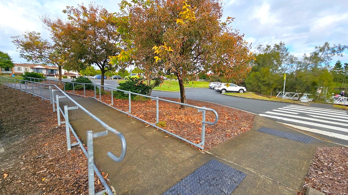 A concrete path with metal railings runs diagonally from the foreground to the left, bordered by mulch and fallen leaves. Trees with autumn foliage line the path. In the background, a road with a zebra crossing is visible, with several parked cars on the right. Houses are on the left, and a grassy area with more trees is on the right. The sky is overcast. A concrete path with metal railings runs diagonally from the foreground to the left, bordered by mulch and fallen leaves. Trees with autumn foliage line the path. In the background, a road with a zebra crossing is visible, with several parked cars on the right. Houses are on the left, and a grassy area with more trees is on the right. The sky is overcast.