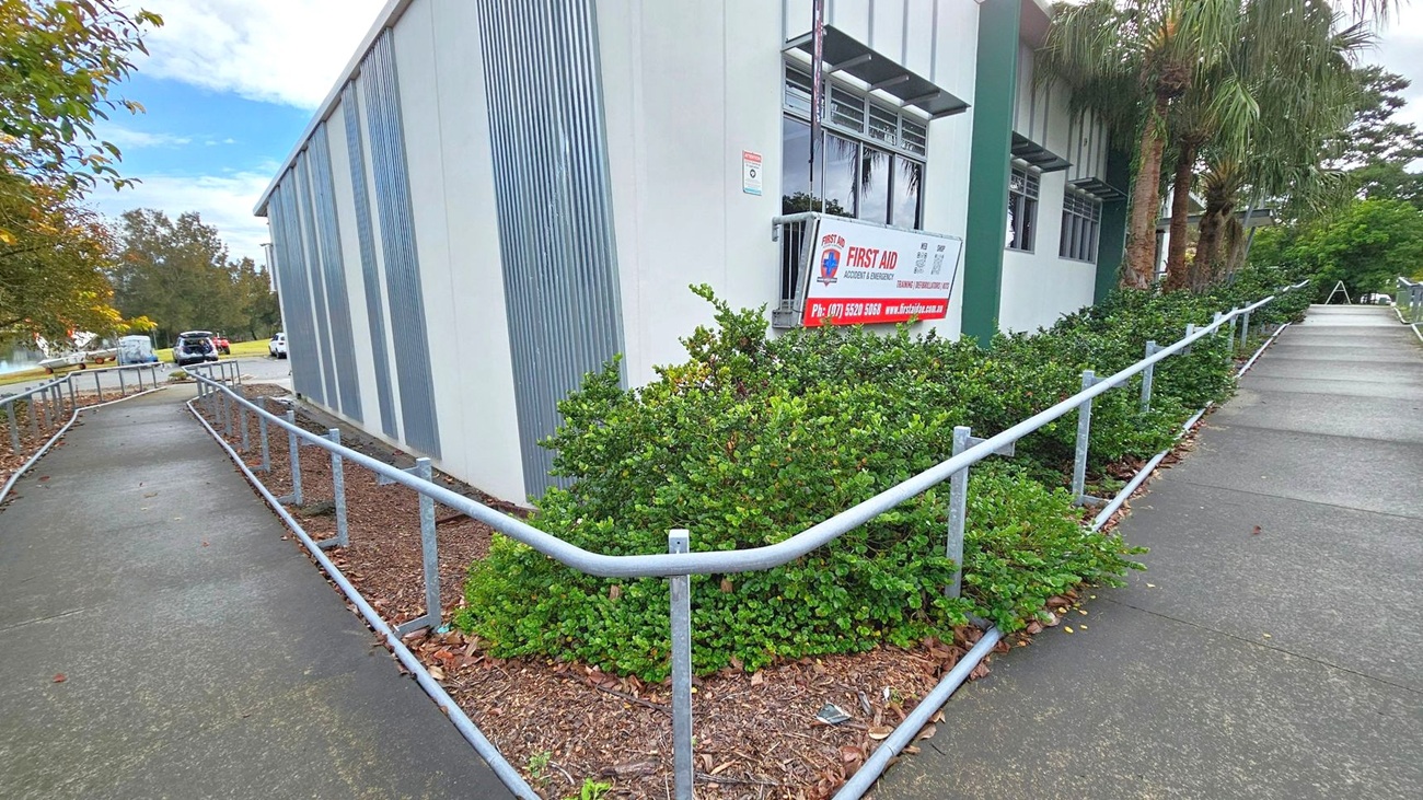 A building with a corrugated metal exterior on the left, featuring a sign for first aid services. A concrete path with metal railings runs alongside the building, bordered by green shrubs. In the background, there are trees and a few parked vehicles. The path leads to a more open area with additional greenery and trees. A building with a corrugated metal exterior on the left, featuring a sign for first aid services. A concrete path with metal railings runs alongside the building, bordered by green shrubs. In the background, there are trees and a few parked vehicles. The path leads to a more open area with additional greenery and trees.