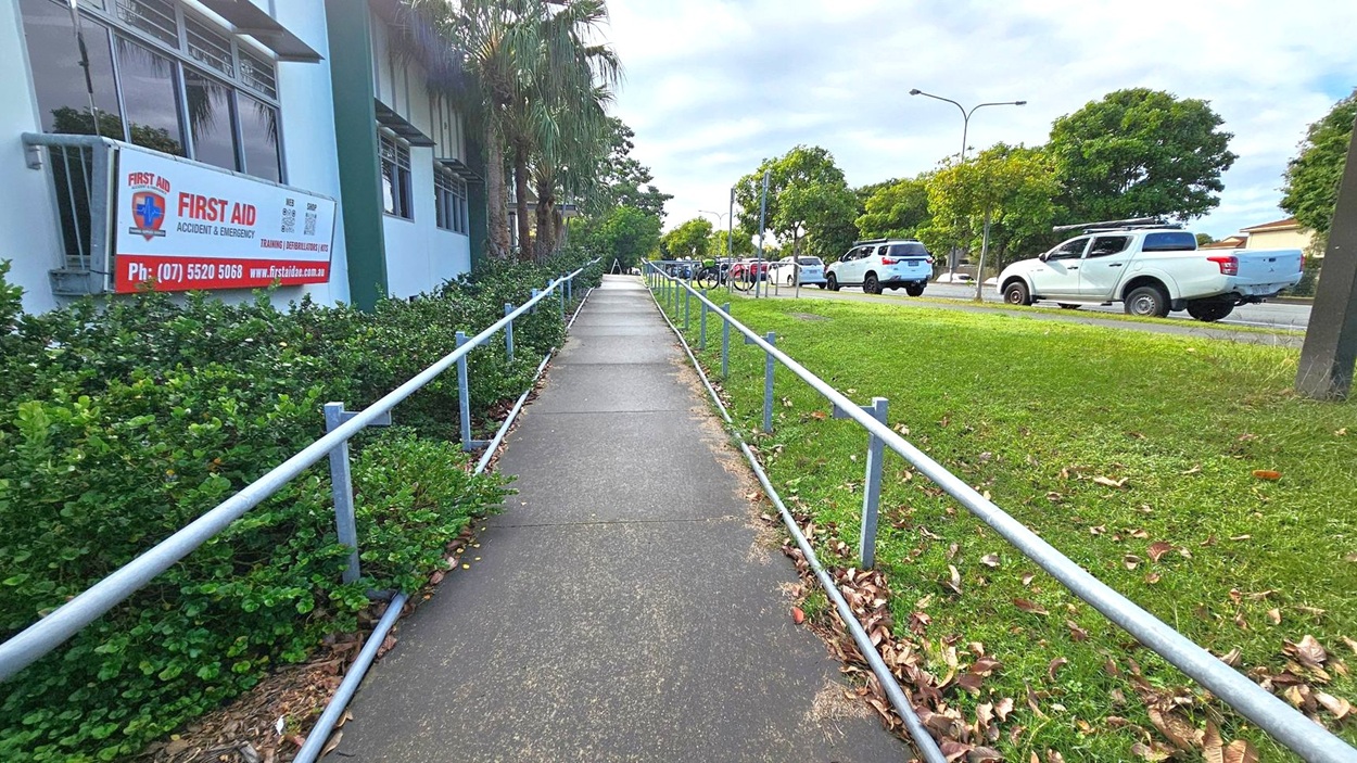 A concrete footpath runs through the centre, bordered by metal railings on both sides. On the left, there's a building with a sign for "First Aid Accident & Emergency" and some green shrubs. On the right, a grassy area with scattered leaves and a few parked white vehicles along the road. Trees line the background, and the sky is overcast. A concrete footpath runs through the centre, bordered by metal railings on both sides. On the left, there's a building with a sign for "First Aid Accident & Emergency" and some green shrubs. On the right, a grassy area with scattered leaves and a few parked white vehicles along the road. Trees line the background, and the sky is overcast.