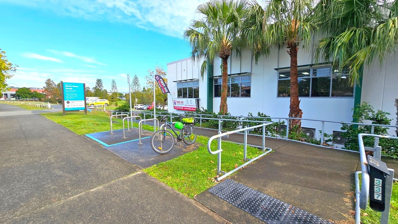 A bicycle with a green helmet is parked at a bike rack on a paved area. To the right, a white building with large windows is partially visible, with palm trees and shrubs in front. A sign for "Sports House Varsity" is on the left, situated on a grassy patch next to a paved path. Several cars are parked in the background, and there are more trees and greenery further back. The ground is a mix of grass and pavement. A bicycle with a green helmet is parked at a bike rack on a paved area. To the right, a white building with large windows is partially visible, with palm trees and shrubs in front. A sign for "Sports House Varsity" is on the left, situated on a grassy patch next to a paved path. Several cars are parked in the background, and there are more trees and greenery further back. The ground is a mix of grass and pavement.