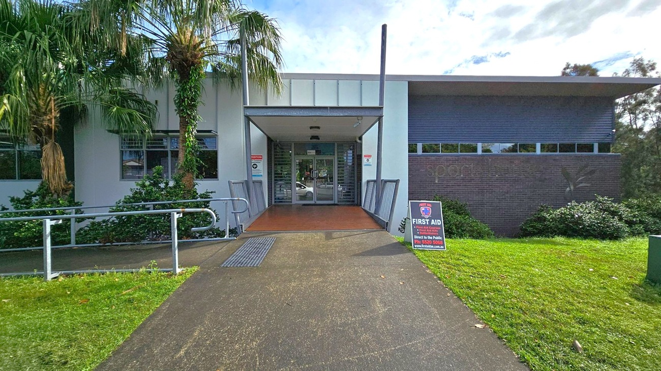 A building entrance with a covered walkway in the centre, flanked by two metal railings. The building has a modern design with a mix of white and dark grey walls. On the left, there are palm trees and shrubs near the windows. On the right, a signboard for first aid services is placed on the grass. The path leading to the entrance is a mix of concrete and grass. The ceiling of the entrance is flat and features a light fixture. A building entrance with a covered walkway in the centre, flanked by two metal railings. The building has a modern design with a mix of white and dark grey walls. On the left, there are palm trees and shrubs near the windows. On the right, a signboard for first aid services is placed on the grass. The path leading to the entrance is a mix of concrete and grass. The ceiling of the entrance is flat and features a light fixture.