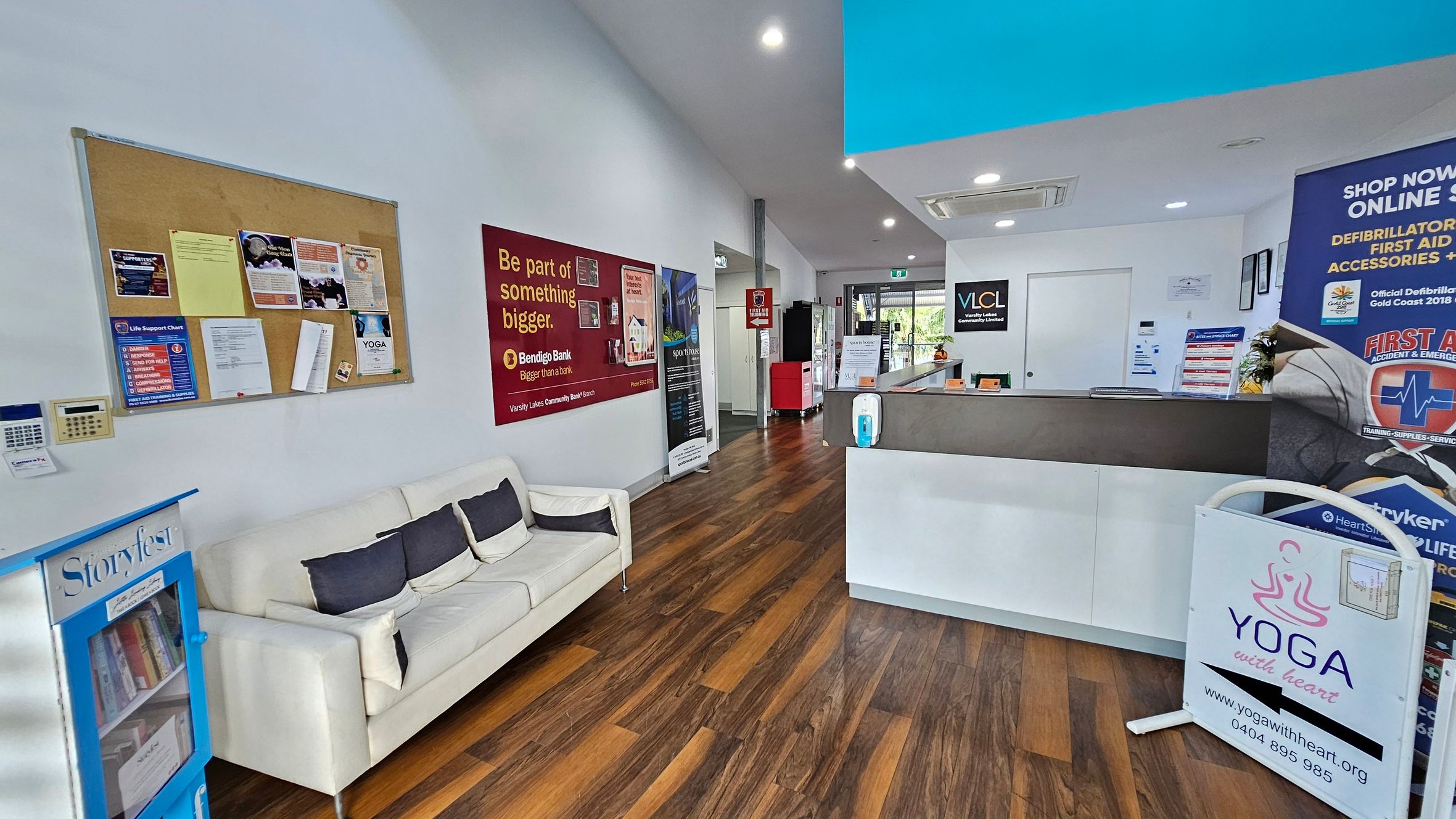 A reception area with wooden flooring and a white ceiling. On the left, a white sofa with dark cushions is placed beneath a bulletin board filled with various notices. A small bookshelf labelled "Storyfest" is nearby. On the right, a reception desk with promotional materials and a sign for "Yoga with Heart" is visible. The back wall features a door and a window, with more posters and a red vending machine. Bright lighting illuminates the space. A reception area with wooden flooring and a white ceiling. On the left, a white sofa with dark cushions is placed beneath a bulletin board filled with various notices. A small bookshelf labelled "Storyfest" is nearby. On the right, a reception desk with promotional materials and a sign for "Yoga with Heart" is visible. The back wall features a door and a window, with more posters and a red vending machine. Bright lighting illuminates the space.