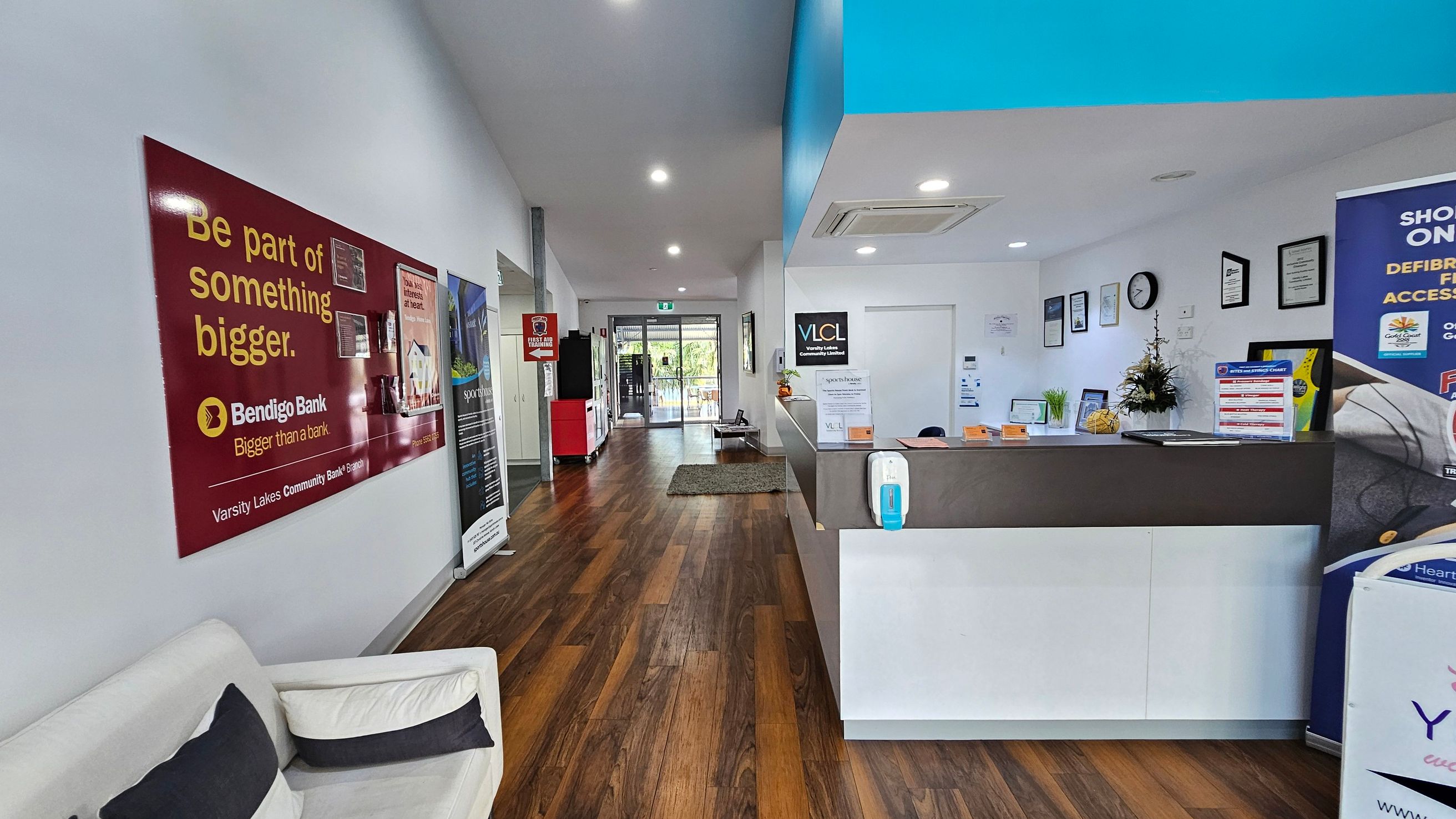 A reception area with a wooden floor and white walls. On the left, a red Bendigo Bank poster is displayed above a white sofa with black and white cushions. The right side features a reception desk with a hand sanitiser dispenser and various informational signs. Behind the desk, framed certificates and a clock are mounted on the wall. The background shows a glass door leading outside, with a small green mat on the floor. The ceiling has recessed lighting. A reception area with a wooden floor and white walls. On the left, a red Bendigo Bank poster is displayed above a white sofa with black and white cushions. The right side features a reception desk with a hand sanitiser dispenser and various informational signs. Behind the desk, framed certificates and a clock are mounted on the wall. The background shows a glass door leading outside, with a small green mat on the floor. The ceiling has recessed lighting.