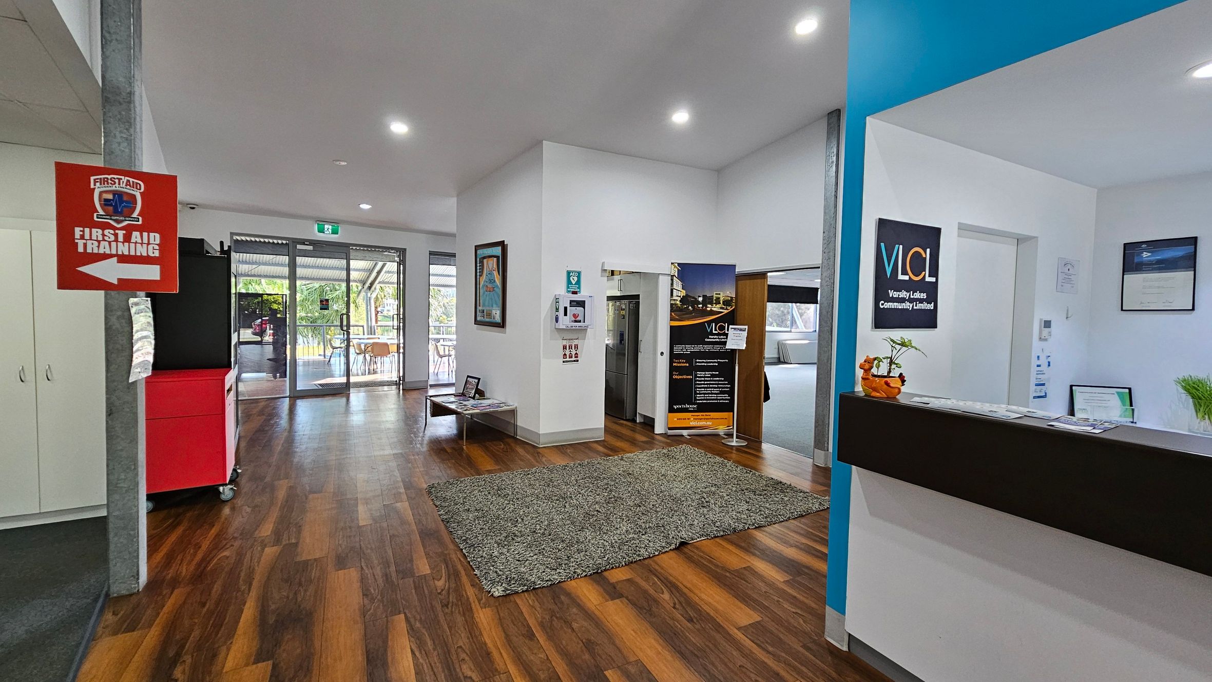 A reception area with a wooden floor and white ceiling. On the left, a red sign indicates "First Aid Training" with an arrow. A red cabinet and white cupboards are nearby. In the centre, a green rug lies on the floor, with a table and posters on the wall. Glass doors lead outside. On the right, a reception desk displays a sign for Varsity Lakes Community Limited, with plants and documents on top. A blue wall section adds contrast. A reception area with a wooden floor and white ceiling. On the left, a red sign indicates "First Aid Training" with an arrow. A red cabinet and white cupboards are nearby. In the centre, a green rug lies on the floor, with a table and posters on the wall. Glass doors lead outside. On the right, a reception desk displays a sign for Varsity Lakes Community Limited, with plants and documents on top. A blue wall section adds contrast.