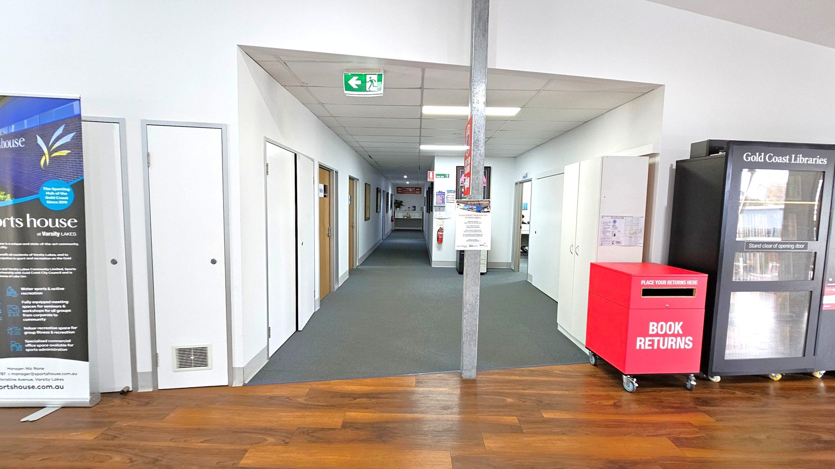 A hallway with a wooden floor leading to a carpeted area. On the left, there's a large banner with text and graphics. Several doors line the left wall. On the right, there's a red book return box on wheels and a black library vending machine labelled "Gold Coast Libraries." The ceiling has fluorescent lights and an exit sign. A fire extinguisher is mounted on the wall in the distance. A hallway with a wooden floor leading to a carpeted area. On the left, there's a large banner with text and graphics. Several doors line the left wall. On the right, there's a red book return box on wheels and a black library vending machine labelled "Gold Coast Libraries." The ceiling has fluorescent lights and an exit sign. A fire extinguisher is mounted on the wall in the distance.