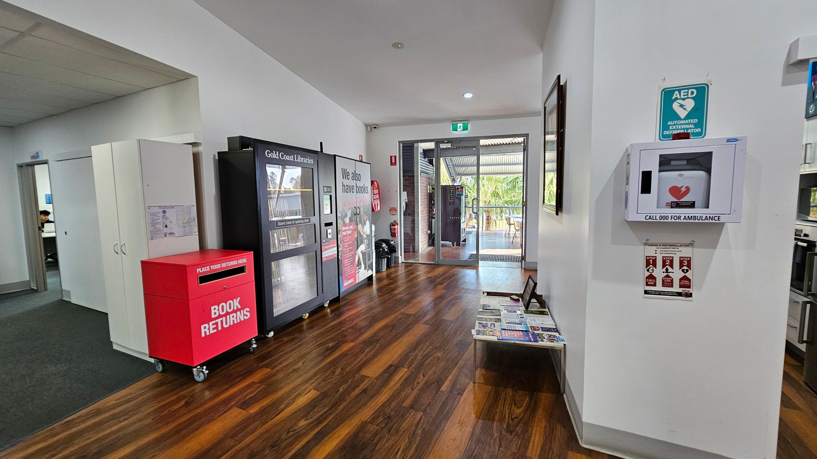 A library entrance area with a wooden floor. On the left, there's a red book return bin and a vending machine for library items. A doorway leads to another room. On the right, a wall-mounted automated external defibrillator (AED) is visible with instructions below. In the centre, glass doors open to an outdoor area with tables and chairs. A small table with brochures is against the right wall. The ceiling is white with recessed lighting. A library entrance area with a wooden floor. On the left, there's a red book return bin and a vending machine for library items. A doorway leads to another room. On the right, a wall-mounted automated external defibrillator (AED) is visible with instructions below. In the centre, glass doors open to an outdoor area with tables and chairs. A small table with brochures is against the right wall. The ceiling is white with recessed lighting.