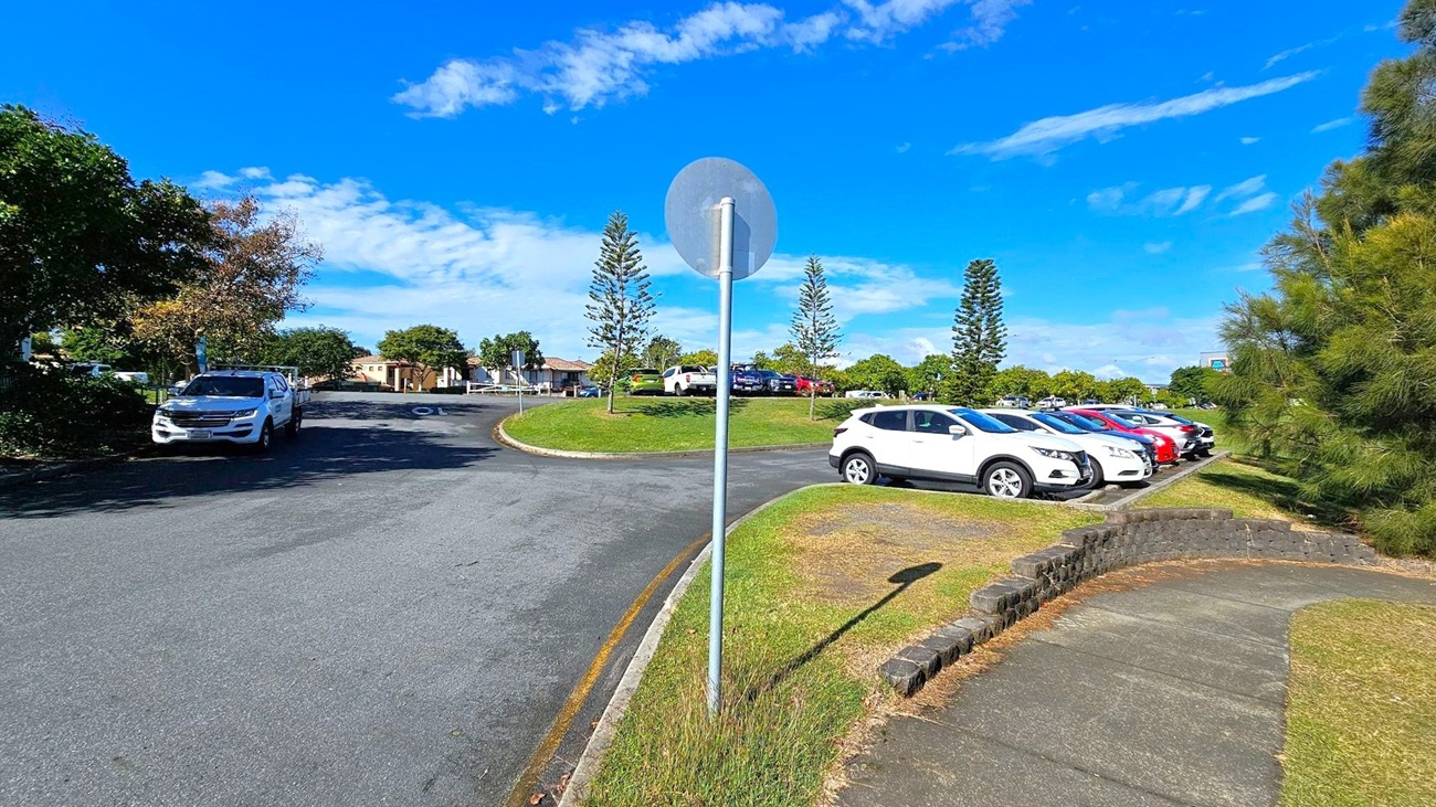 A curved road with a white vehicle parked on the left side. In the centre, a metal pole with a circular sign. On the right, a row of parked cars in various colours, including white and red, on a grassy area. Trees and shrubs are scattered throughout, with a few tall, narrow trees in the background. A paved footpath runs alongside the grass, bordered by a low brick wall. A curved road with a white vehicle parked on the left side. In the centre, a metal pole with a circular sign. On the right, a row of parked cars in various colours, including white and red, on a grassy area. Trees and shrubs are scattered throughout, with a few tall, narrow trees in the background. A paved footpath runs alongside the grass, bordered by a low brick wall.