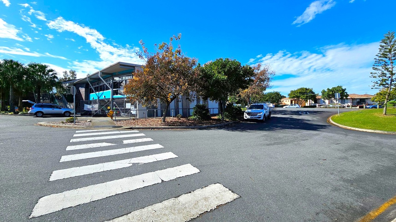 A zebra crossing leads to a building with a modern design on the left, featuring large windows and a slanted roof. Several trees with autumn foliage are in front of the building. A silver car is parked nearby. On the right, a white vehicle is parked along a curved road, with a grassy area and more trees. In the background, there are residential houses with tiled roofs. The sky is clear with a few clouds. A zebra crossing leads to a building with a modern design on the left, featuring large windows and a slanted roof. Several trees with autumn foliage are in front of the building. A silver car is parked nearby. On the right, a white vehicle is parked along a curved road, with a grassy area and more trees. In the background, there are residential houses with tiled roofs. The sky is clear with a few clouds.