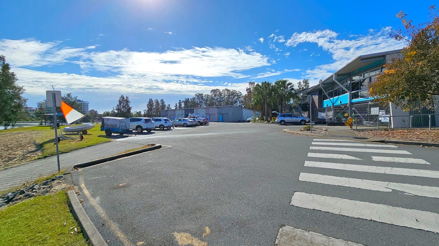 A car park with several vehicles is in the centre, surrounded by trees and a building on the right with a blue facade. A pedestrian crossing is visible in the foreground. On the left, a grassy area with a small boat and a trailer is near a path. A sign with an orange and white flag is also on the left. The background features more trees and a few buildings. A car park with several vehicles is in the centre, surrounded by trees and a building on the right with a blue facade. A pedestrian crossing is visible in the foreground. On the left, a grassy area with a small boat and a trailer is near a path. A sign with an orange and white flag is also on the left. The background features more trees and a few buildings.
