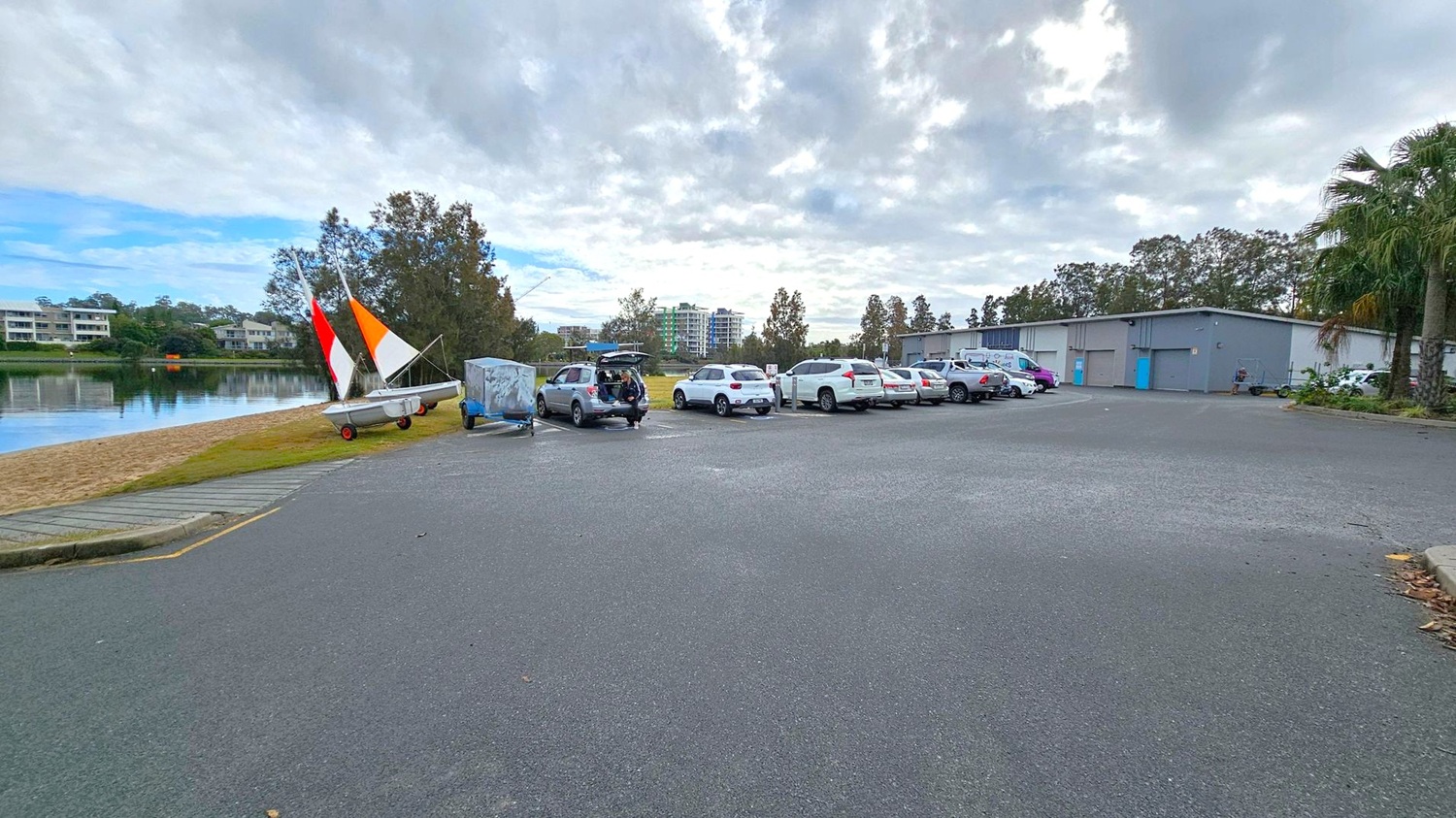 A car park with several vehicles is on the right, with a row of white and silver cars. In the middle, a car has its boot open. On the left, a small sailboat with red, orange, and white sails is on a grassy area near a sandy shore. In the background, there are trees and a few multi-storey buildings. A large grey building with blue doors is on the right. The ground is paved, and there are some palm trees on the far right. A car park with several vehicles is on the right, with a row of white and silver cars. In the middle, a car has its boot open. On the left, a small sailboat with red, orange, and white sails is on a grassy area near a sandy shore. In the background, there are trees and a few multi-storey buildings. A large grey building with blue doors is on the right. The ground is paved, and there are some palm trees on the far right.