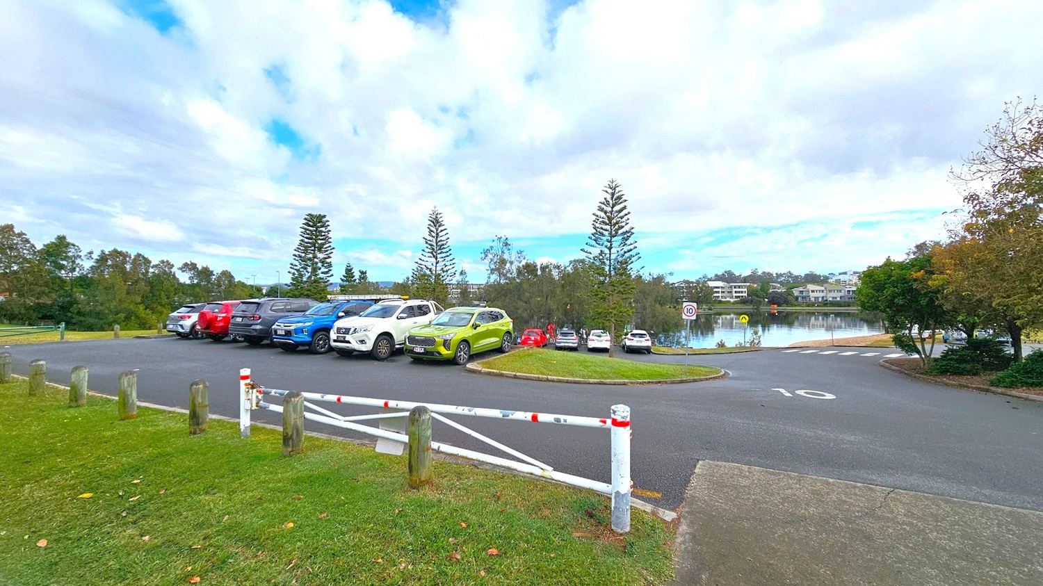 A car park with several vehicles, including a bright green car, is on the left. The area is bordered by wooden posts and a white metal gate with red markings. In the background, there are tall trees and a body of water with buildings visible on the far side. The road in the foreground has a speed limit sign marked "10". The scene is surrounded by greenery, with trees on the right. A car park with several vehicles, including a bright green car, is on the left. The area is bordered by wooden posts and a white metal gate with red markings. In the background, there are tall trees and a body of water with buildings visible on the far side. The road in the foreground has a speed limit sign marked "10". The scene is surrounded by greenery, with trees on the right.