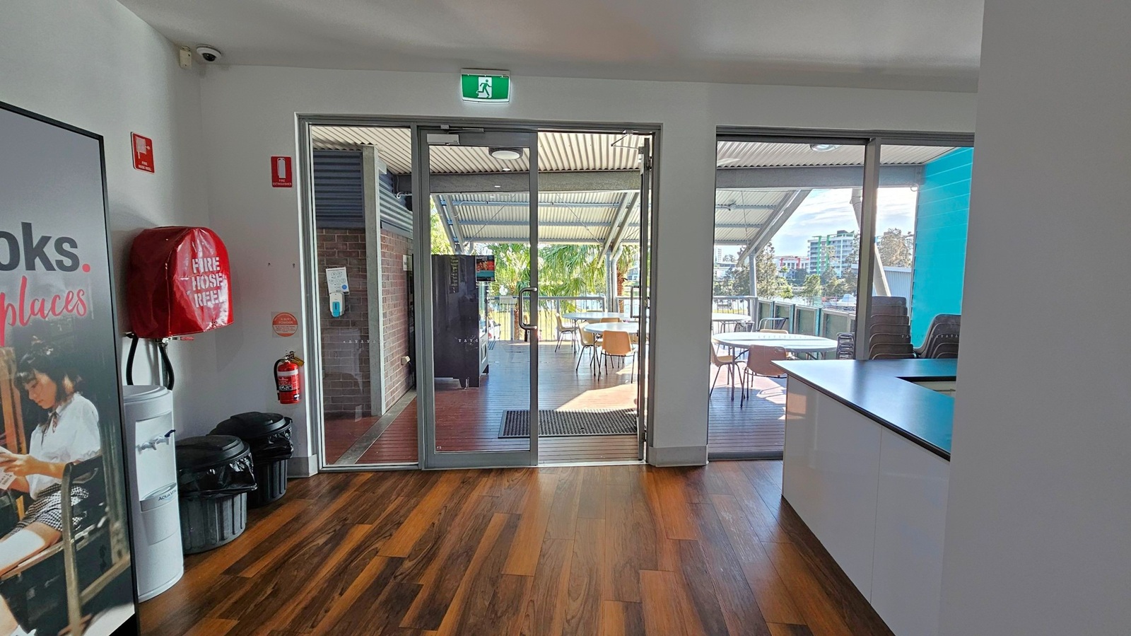 A room with wooden flooring and a white ceiling. On the left, there's a poster, a fire hose reel, a water cooler, and two bins. In the centre, glass doors lead to an outdoor area with tables and chairs under a metal roof. A fire extinguisher is mounted on the wall near the door. On the right, there's a countertop with a glossy finish. Outside, trees and buildings are visible in the background. A room with wooden flooring and a white ceiling. On the left, there's a poster, a fire hose reel, a water cooler, and two bins. In the centre, glass doors lead to an outdoor area with tables and chairs under a metal roof. A fire extinguisher is mounted on the wall near the door. On the right, there's a countertop with a glossy finish. Outside, trees and buildings are visible in the background.