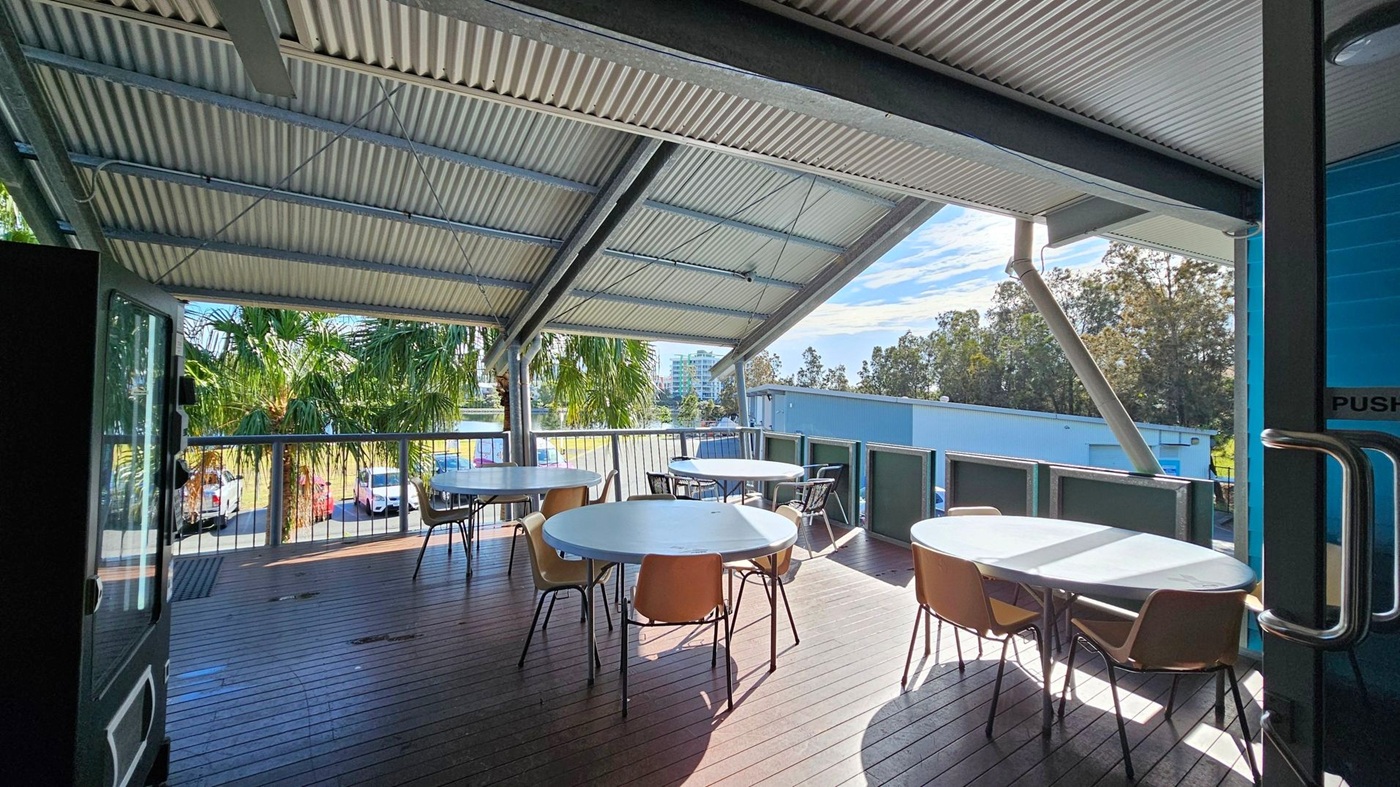A covered outdoor deck with a corrugated metal roof and wooden flooring. Several round tables with chairs are arranged on the deck. The area is surrounded by a railing, and palm trees are visible in the background. Beyond the trees, there are parked cars and a glimpse of buildings. A glass door with a metal handle is partially open on the right side. A covered outdoor deck with a corrugated metal roof and wooden flooring. Several round tables with chairs are arranged on the deck. The area is surrounded by a railing, and palm trees are visible in the background. Beyond the trees, there are parked cars and a glimpse of buildings. A glass door with a metal handle is partially open on the right side.
