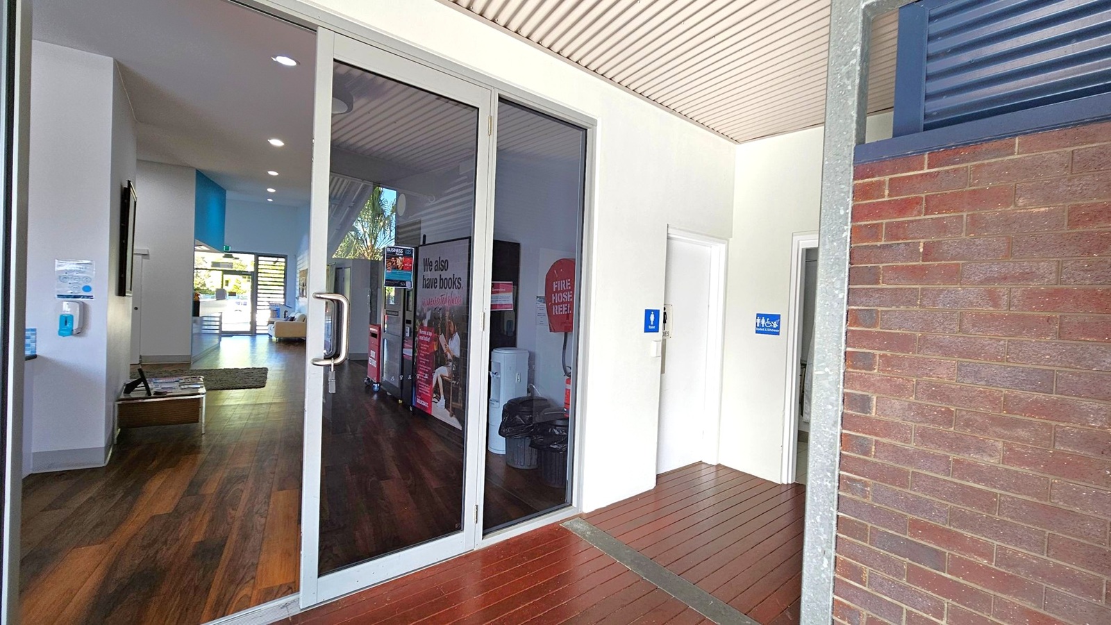 On the left, there is a glass door with a metal handle leading into a room with wooden flooring. Inside, a wall-mounted screen and a hand sanitiser dispenser are visible. In the middle, a sign reads "We also have books" next to a fire hose reel. On the right, there are two doors with blue signs indicating restrooms. The floor outside the doors is wooden, and the ceiling has a light-coloured, ribbed design. A brick wall is partially visible on the far right. On the left, there is a glass door with a metal handle leading into a room with wooden flooring. Inside, a wall-mounted screen and a hand sanitiser dispenser are visible. In the middle, a sign reads "We also have books" next to a fire hose reel. On the right, there are two doors with blue signs indicating restrooms. The floor outside the doors is wooden, and the ceiling has a light-coloured, ribbed design. A brick wall is partially visible on the far right.