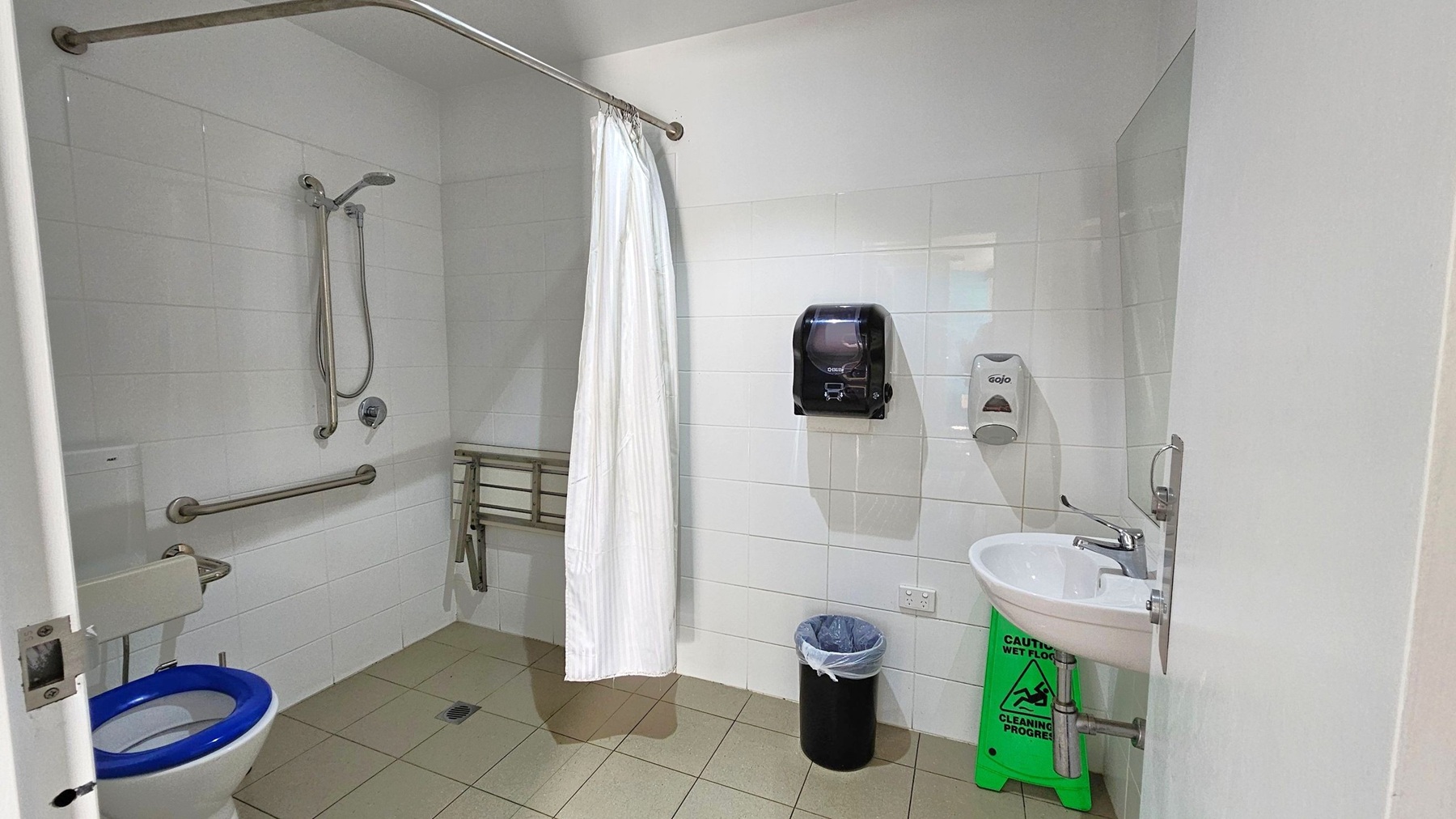 Accessible bathroom with white tiled walls and a light green tiled floor. On the left, a toilet with a blue seat and support bars. A shower area with a handheld showerhead and a white curtain. In the centre, a foldable shower seat. On the right, a sink with a soap dispenser and a paper towel dispenser above a bin. A green "Caution: Wet Floor" sign is beneath the sink. The ceiling is plain white. Accessible bathroom with white tiled walls and a light green tiled floor. On the left, a toilet with a blue seat and support bars. A shower area with a handheld showerhead and a white curtain. In the centre, a foldable shower seat. On the right, a sink with a soap dispenser and a paper towel dispenser above a bin. A green "Caution: Wet Floor" sign is beneath the sink. The ceiling is plain white.
