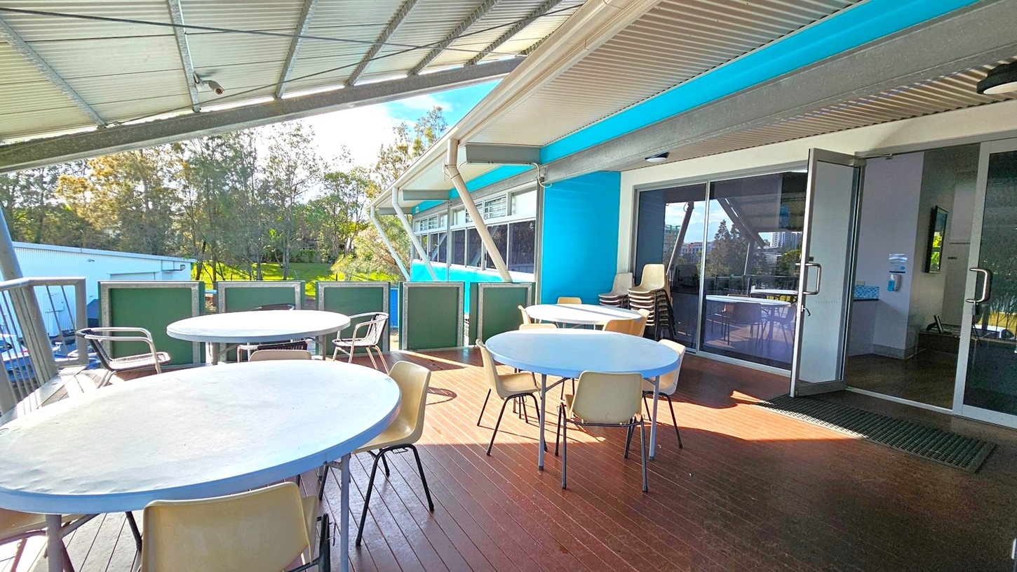 A covered outdoor seating area with several round tables and chairs on a wooden deck. On the right, there are glass doors leading inside a building with a blue exterior. The ceiling is metal, and the area is partially enclosed with green panels. In the background, there are trees and a grassy area, with a white building visible to the left. A covered outdoor seating area with several round tables and chairs on a wooden deck. On the right, there are glass doors leading inside a building with a blue exterior. The ceiling is metal, and the area is partially enclosed with green panels. In the background, there are trees and a grassy area, with a white building visible to the left.