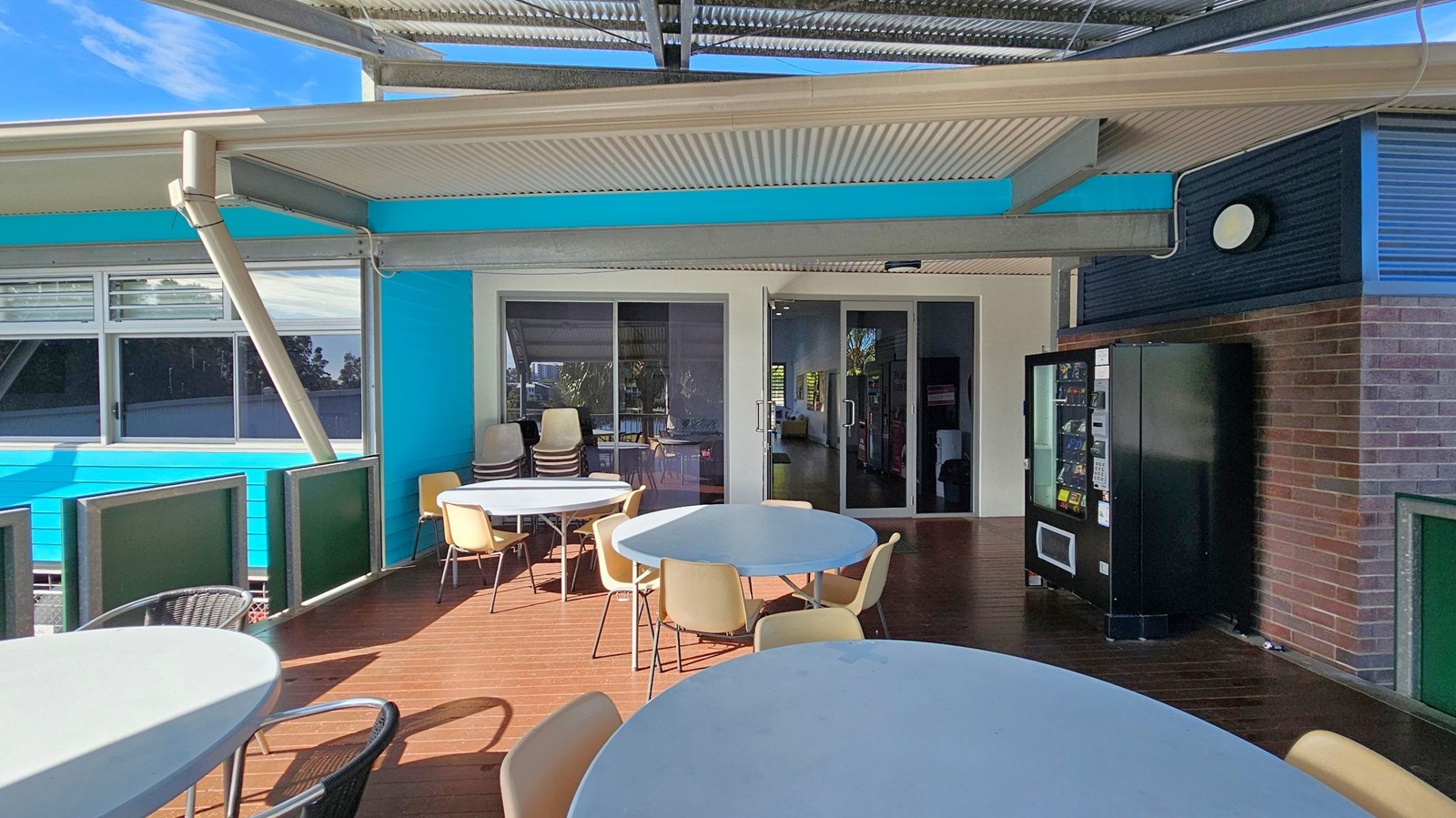 Outdoor seating area with round tables and chairs on a wooden deck. On the right, a black vending machine is positioned against a brick wall. The background features a blue and white building with large windows and a glass door. The ceiling is made of corrugated metal, supported by beams. A stack of chairs is visible near the door. Outdoor seating area with round tables and chairs on a wooden deck. On the right, a black vending machine is positioned against a brick wall. The background features a blue and white building with large windows and a glass door. The ceiling is made of corrugated metal, supported by beams. A stack of chairs is visible near the door.