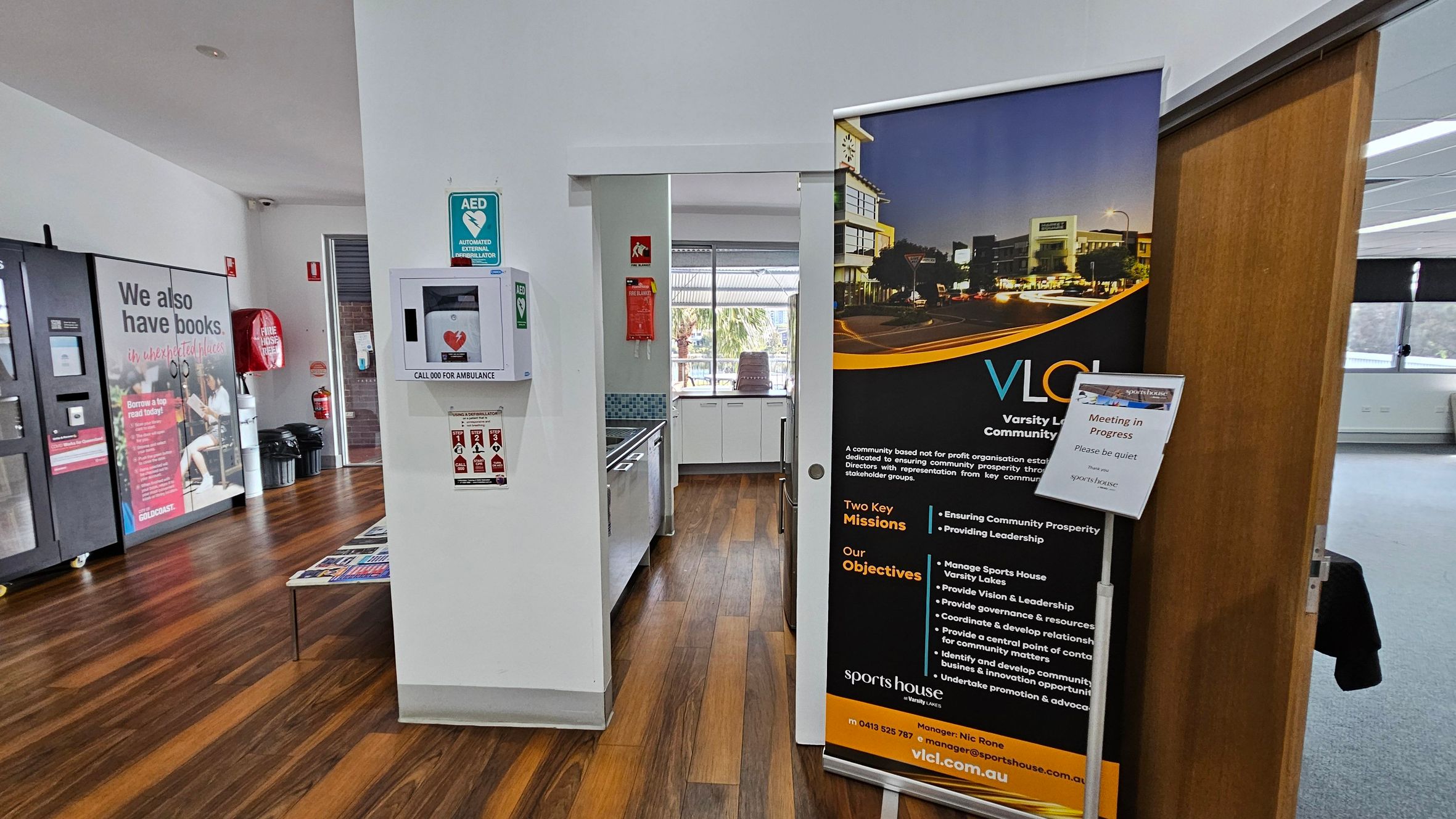 A room with wooden flooring and white walls. On the left, a large poster with text about books and a vending machine. In the centre, an AED mounted on the wall and a small kitchen area with a sink and window. On the right, a large banner with information about Varsity Lakes Community Limited and a sign indicating a meeting in progress. A partially open wooden door leads to a carpeted area. A room with wooden flooring and white walls. On the left, a large poster with text about books and a vending machine. In the centre, an AED mounted on the wall and a small kitchen area with a sink and window. On the right, a large banner with information about Varsity Lakes Community Limited and a sign indicating a meeting in progress. A partially open wooden door leads to a carpeted area.