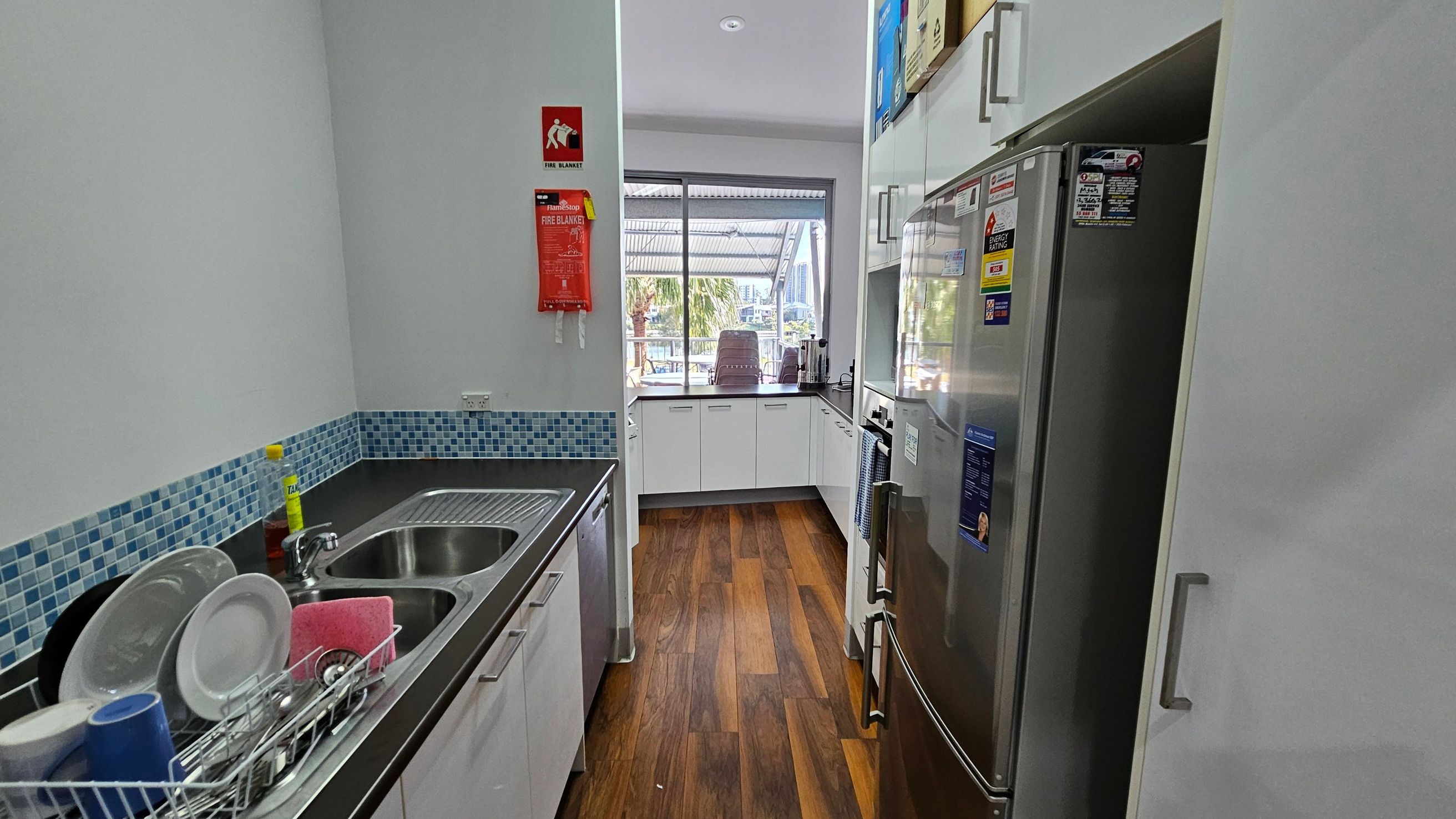 A narrow kitchen with a wooden floor. On the left, there's a countertop with a double sink, a dish rack with plates and cups, and a bottle of dish soap. Above the sink is a blue mosaic tile backsplash. On the right, a stainless steel refrigerator with various stickers. In the centre, a window with a view of outdoor seating and palm trees. A fire blanket is mounted on the wall near the window. White cabinets line the walls, and the ceiling has recessed lighting. A narrow kitchen with a wooden floor. On the left, there's a countertop with a double sink, a dish rack with plates and cups, and a bottle of dish soap. Above the sink is a blue mosaic tile backsplash. On the right, a stainless steel refrigerator with various stickers. In the centre, a window with a view of outdoor seating and palm trees. A fire blanket is mounted on the wall near the window. White cabinets line the walls, and the ceiling has recessed lighting.