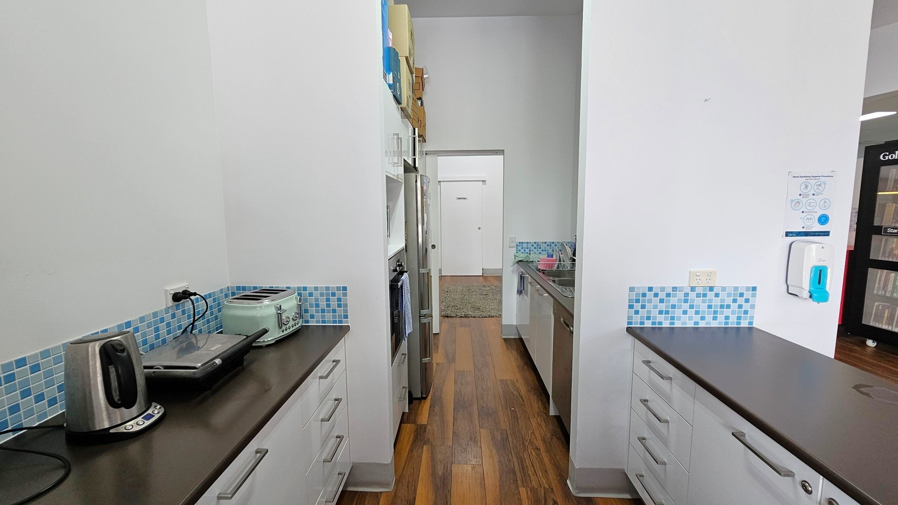 A narrow kitchen with wooden flooring and white walls. On the left, a black countertop holds a stainless steel kettle, a sandwich press, and a mint green toaster. Above are blue mosaic tiles. On the right, a similar countertop with a sink and dish rack. A hand sanitiser dispenser and a poster are mounted on the wall. In the background, a doorway leads to a room with a green rug. Shelves and a stainless steel fridge are visible along the left wall. A narrow kitchen with wooden flooring and white walls. On the left, a black countertop holds a stainless steel kettle, a sandwich press, and a mint green toaster. Above are blue mosaic tiles. On the right, a similar countertop with a sink and dish rack. A hand sanitiser dispenser and a poster are mounted on the wall. In the background, a doorway leads to a room with a green rug. Shelves and a stainless steel fridge are visible along the left wall.