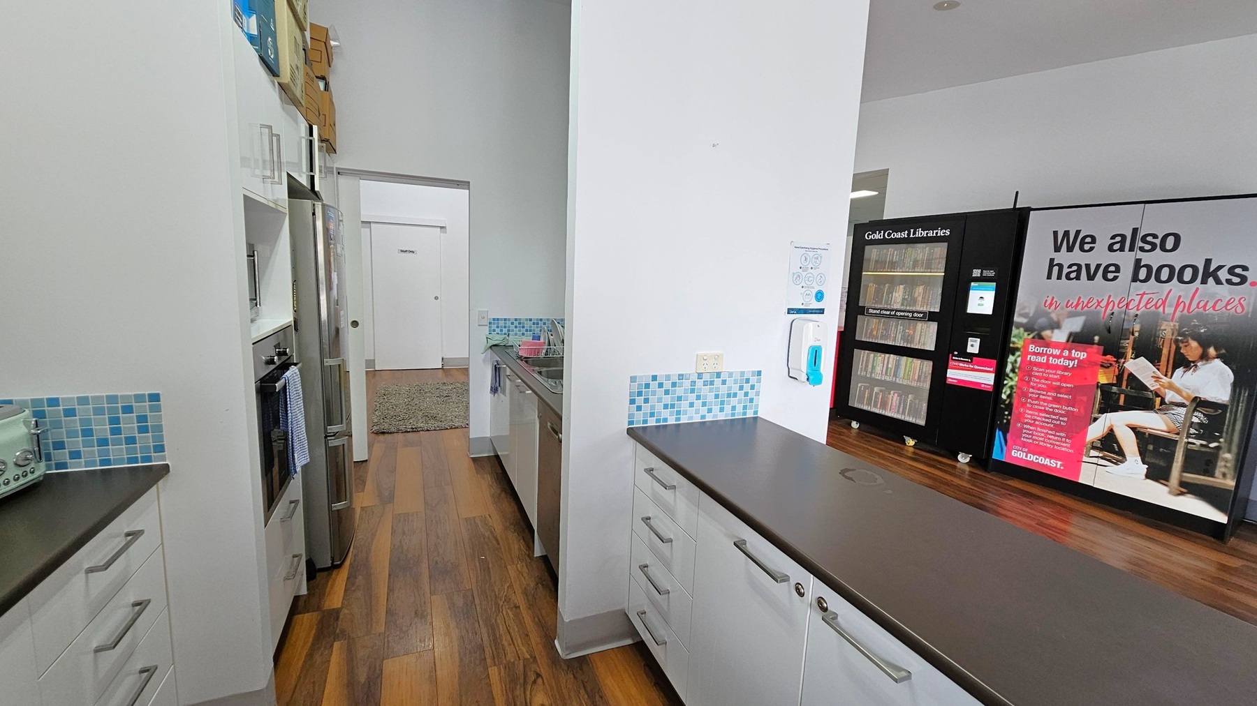 A modern kitchen area with white cabinets and black countertops. On the left, there is a microwave and a fridge, with a blue and white checkered towel hanging from the oven handle. The floor is wooden. In the background, a green rug leads to a white door. On the right, a vending machine with books and a large poster advertising library services. Blue mosaic tiles are used as a backsplash above the sink and counter. A hand sanitiser dispenser is mounted on the wall. A modern kitchen area with white cabinets and black countertops. On the left, there is a microwave and a fridge, with a blue and white checkered towel hanging from the oven handle. The floor is wooden. In the background, a green rug leads to a white door. On the right, a vending machine with books and a large poster advertising library services. Blue mosaic tiles are used as a backsplash above the sink and counter. A hand sanitiser dispenser is mounted on the wall.