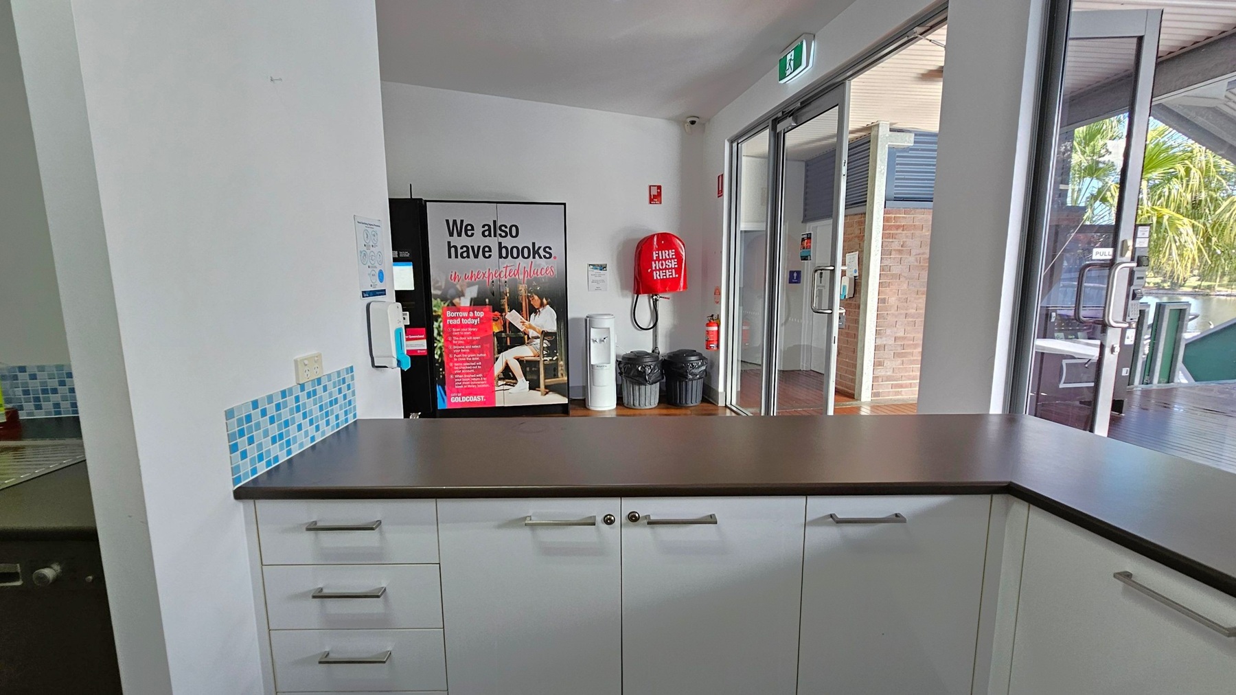 A room with a countertop in the foreground, featuring white cabinets with silver handles. On the left, there's a wall with a blue-tiled backsplash and a sink. In the centre, a poster with text and a fire hose reel are mounted on the wall. To the right, glass doors lead outside, showing a view of a brick wall and palm trees. The ceiling has a white finish, and the floor is wooden. An exit sign is above the doors. A room with a countertop in the foreground, featuring white cabinets with silver handles. On the left, there's a wall with a blue-tiled backsplash and a sink. In the centre, a poster with text and a fire hose reel are mounted on the wall. To the right, glass doors lead outside, showing a view of a brick wall and palm trees. The ceiling has a white finish, and the floor is wooden. An exit sign is above the doors.