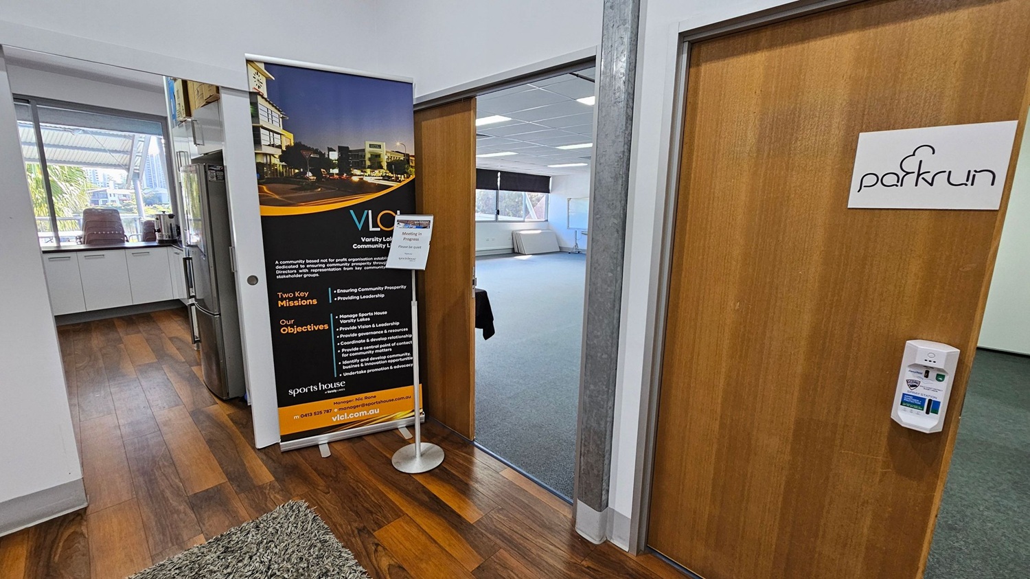 A hallway with wooden flooring and a green rug. On the left, there's a kitchen area with white cabinets, a refrigerator, and a window showing an outdoor view with trees and buildings. In the centre, a large vertical banner with text and images stands next to an open door leading to a room with grey carpet and a window. On the right, a wooden door with a "parkrun" sign and a wall-mounted device is visible. A hallway with wooden flooring and a green rug. On the left, there's a kitchen area with white cabinets, a refrigerator, and a window showing an outdoor view with trees and buildings. In the centre, a large vertical banner with text and images stands next to an open door leading to a room with grey carpet and a window. On the right, a wooden door with a "parkrun" sign and a wall-mounted device is visible.