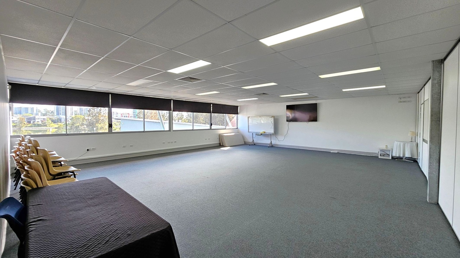 A spacious room with a grey carpeted floor and a grid-patterned ceiling with fluorescent lights. On the left, there are stacked wooden chairs and a table covered with a black cloth. Large windows with black blinds line the left wall, offering a view of buildings and trees outside. On the right, a whiteboard and a wall-mounted TV are visible. A small round table with a white cloth is in the corner. The room is mostly empty, providing an open area. A spacious room with a grey carpeted floor and a grid-patterned ceiling with fluorescent lights. On the left, there are stacked wooden chairs and a table covered with a black cloth. Large windows with black blinds line the left wall, offering a view of buildings and trees outside. On the right, a whiteboard and a wall-mounted TV are visible. A small round table with a white cloth is in the corner. The room is mostly empty, providing an open area.