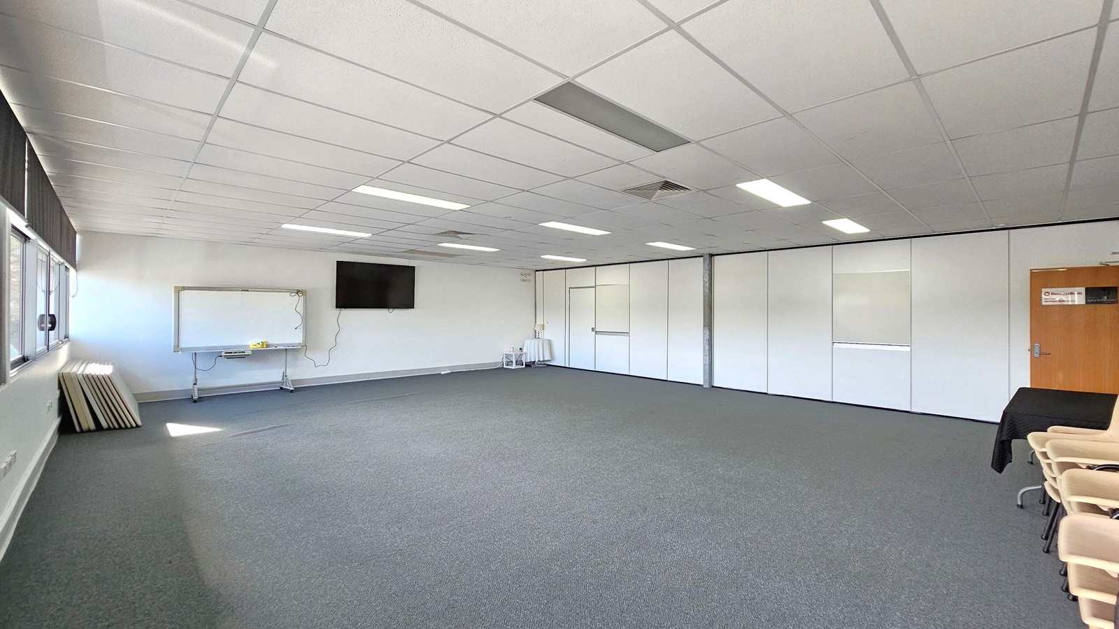 A spacious room with a grey carpeted floor and a white tiled ceiling featuring fluorescent lights. On the left, there are large windows with blinds, a whiteboard, and a wall-mounted TV. The right side has a wooden door and a stack of chairs. The back wall is lined with white partition panels, and a small table with items is visible. A spacious room with a grey carpeted floor and a white tiled ceiling featuring fluorescent lights. On the left, there are large windows with blinds, a whiteboard, and a wall-mounted TV. The right side has a wooden door and a stack of chairs. The back wall is lined with white partition panels, and a small table with items is visible.