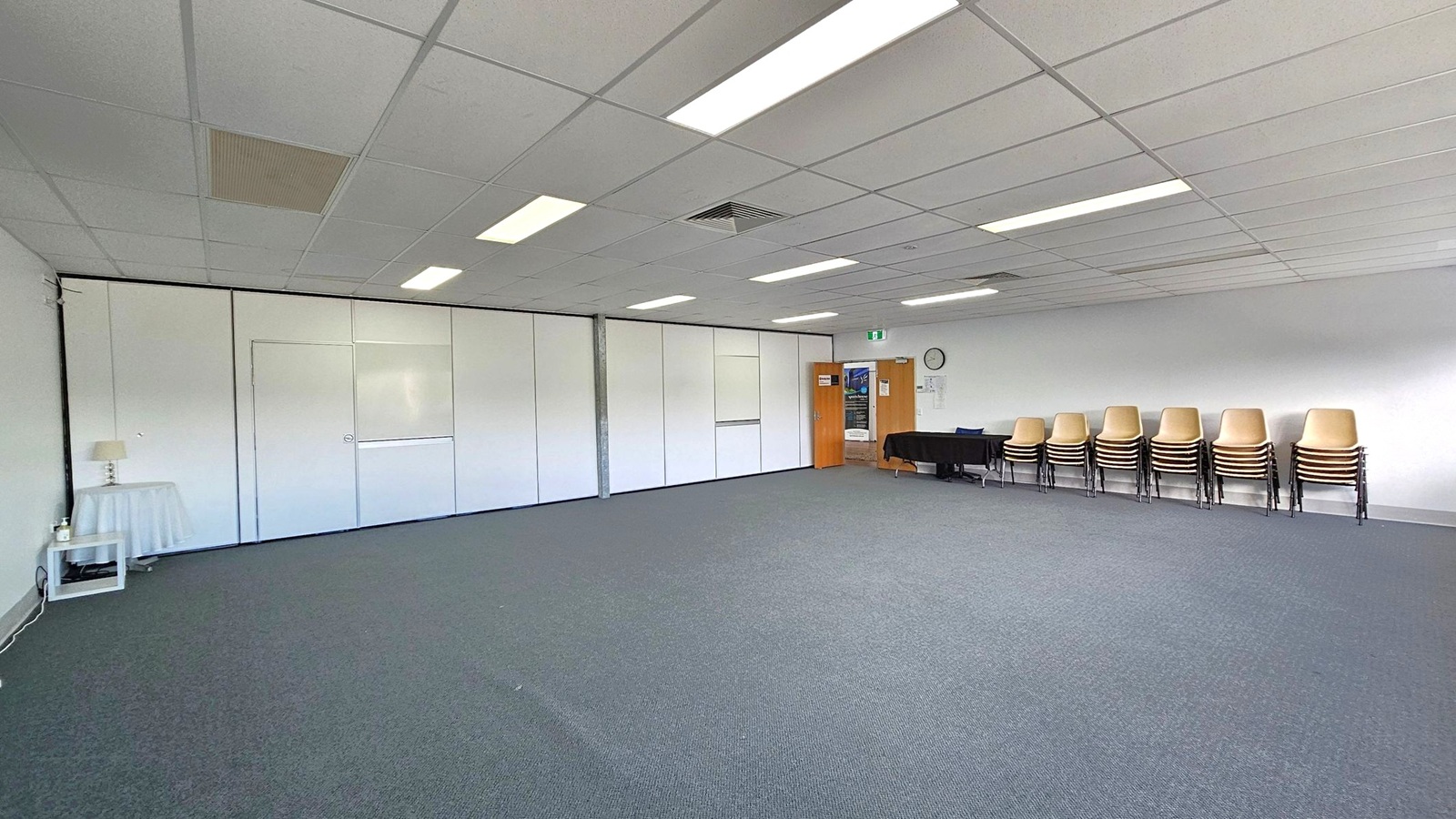A spacious room with a grey carpeted floor and a white tiled ceiling featuring fluorescent lights. On the right, there are stacks of chairs against the wall, next to a black table. A clock is mounted on the wall above. In the background, a wooden door with a poster leads to another area. On the left, a small table with a lamp and a bottle is placed against a partitioned wall. A spacious room with a grey carpeted floor and a white tiled ceiling featuring fluorescent lights. On the right, there are stacks of chairs against the wall, next to a black table. A clock is mounted on the wall above. In the background, a wooden door with a poster leads to another area. On the left, a small table with a lamp and a bottle is placed against a partitioned wall.